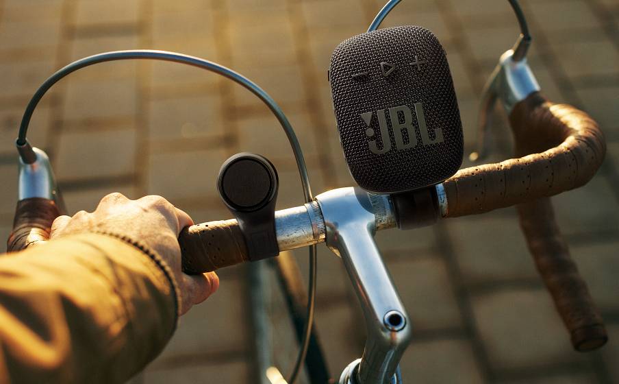 Jedna ruka drží riadidlá bicykla. Prenosný reproduktor je pripevnený na riadidlách. V pozadí je vidieť dláždenú cestu.