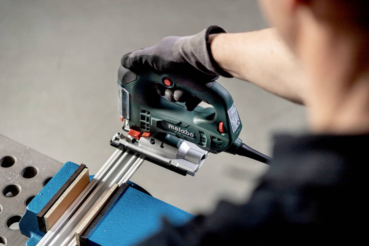 Osoba strihá kovovú konzolu upevnenú vo zveráku pomocou priamočiarej píly. Rukavice chránia ruky.