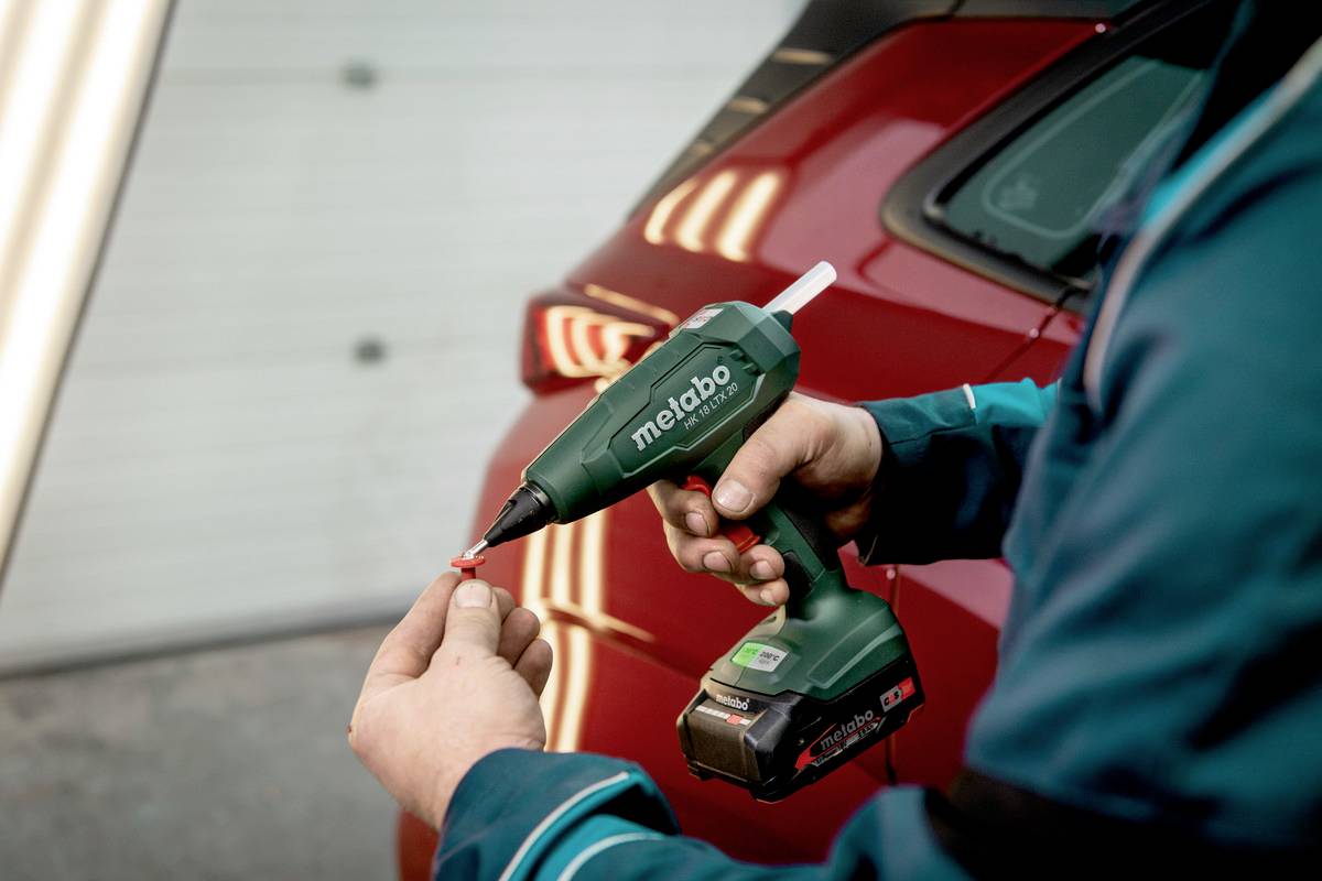 Osoba drží zelený akumulátorový skrutkovač a upevňuje skrutku. V pozadí je vidieť červené auto.