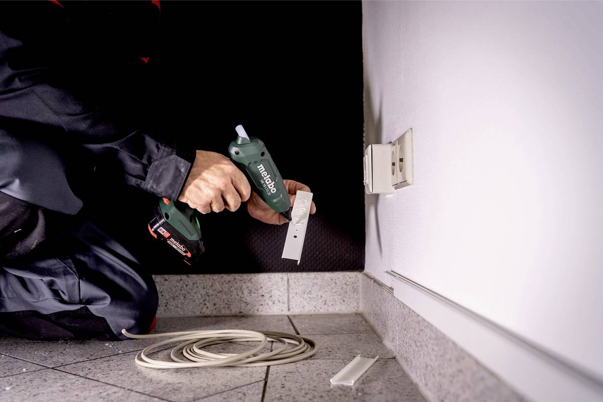Osoba pripevňuje bielu lištu na stenu pomocou vŕtacieho skrutkovača. Kábel leží na podlahe pripravený.