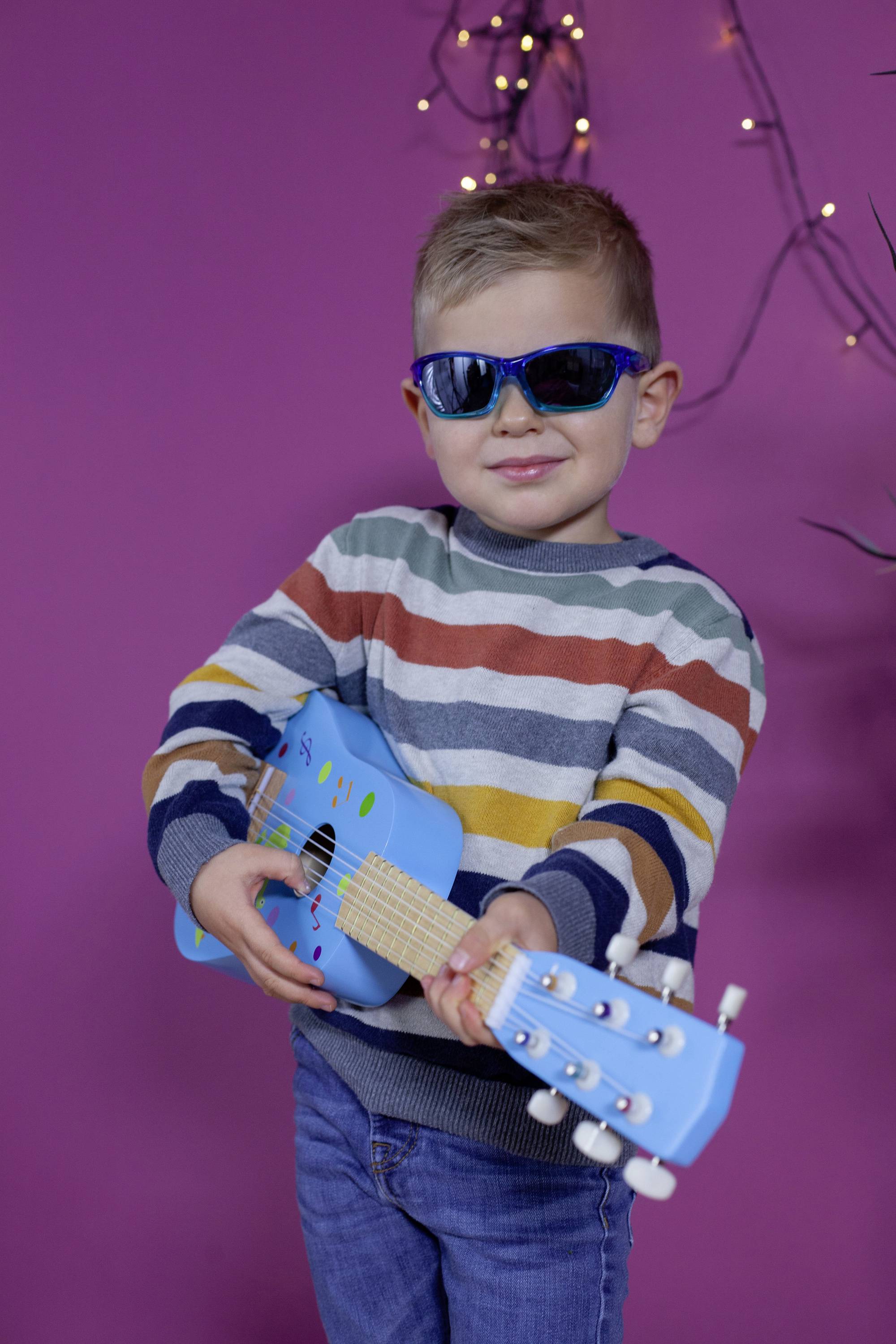 Dieťa s slnečnými okuliarmi hrá na ukulele pred ružovým pozadím. Má na sebe pruhovaný pulóver. V pozadí sú svetelné reťaze.