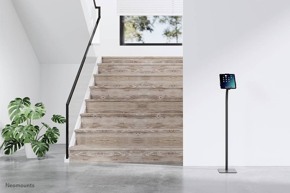 Drevené schodisko v minimalistickom priestore, vedľa ktorého stojí tablet na stojane. Veľká rastlina vľavo.