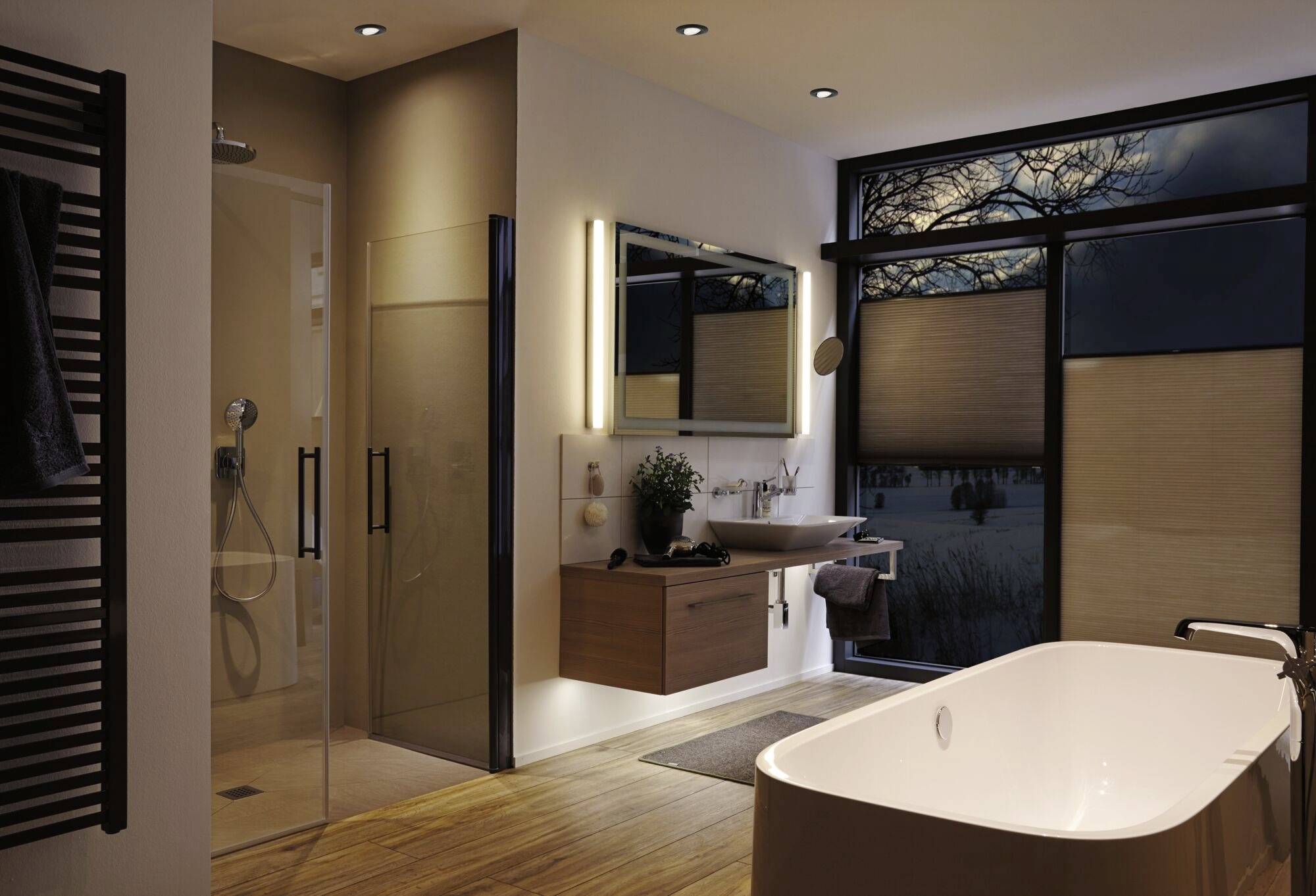 Moderná kúpeľňa so samostatne stojacou vaňou, osvetleným zrkadlom nad umývadlom a veľkou presklenou stenou s výhľadom na stromy.