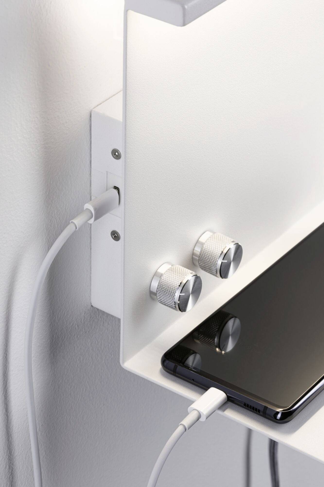 Smartfón sa nabíja na stojanček podobnom nabíjacej ploche; vedľa sú dva strieborné nastavovacie gombíky.