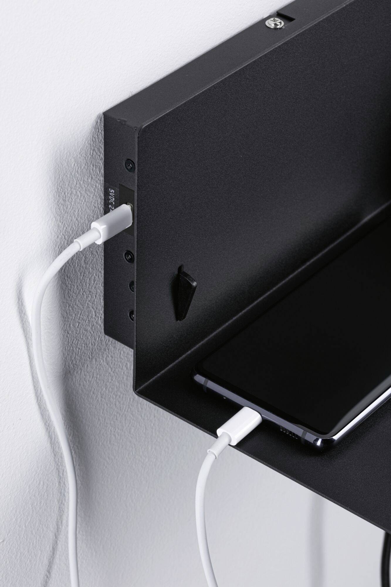 Čierna polica namontovaná na stene s vstavanou USB nabíjačkou. Smartphone sa nabíja pomocou bieleho kábla.