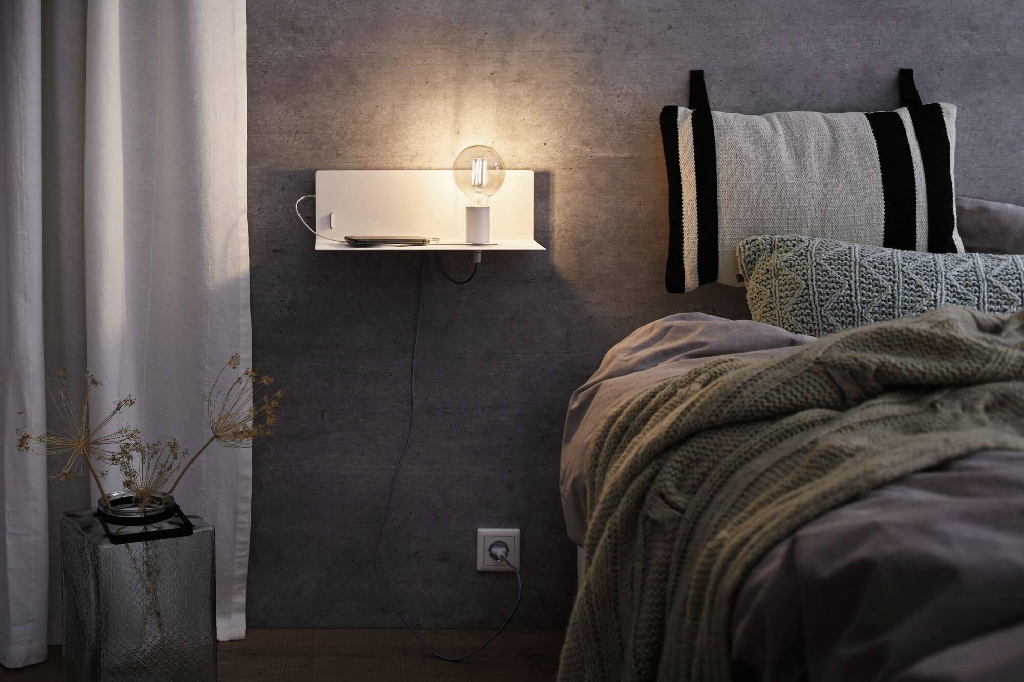 Útulná spálňa so tlmeným svetlom, posteľ s vankúšmi a dekou, nočný stolík s lampou. Moderná, uvoľnená atmosféra.