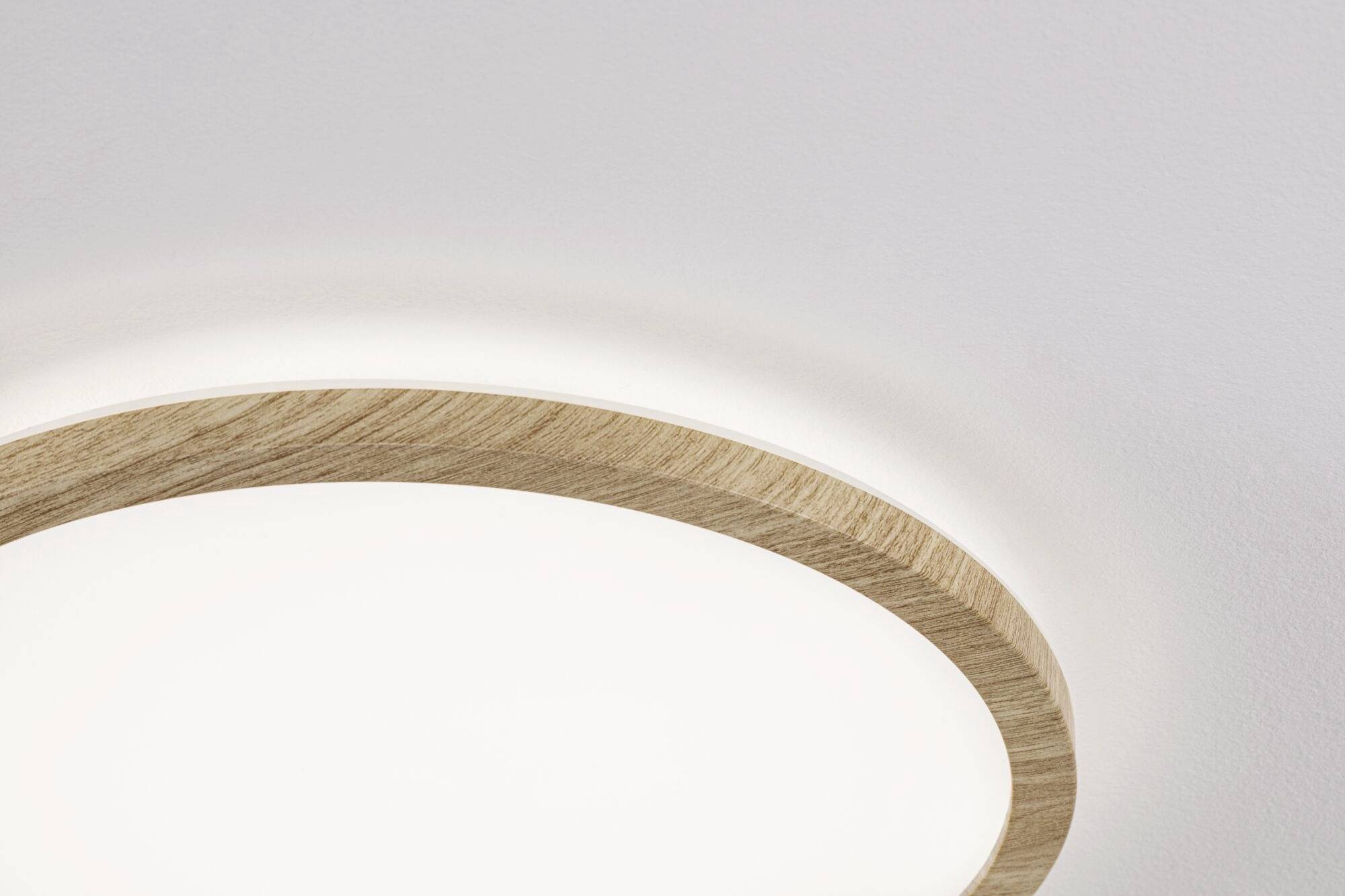 Okruhové stropné svietidlo s dreveným rámom v svetlej miestnosti. Tienidlo žiari jemne a vytvára moderne minimalistickú atmosféru.