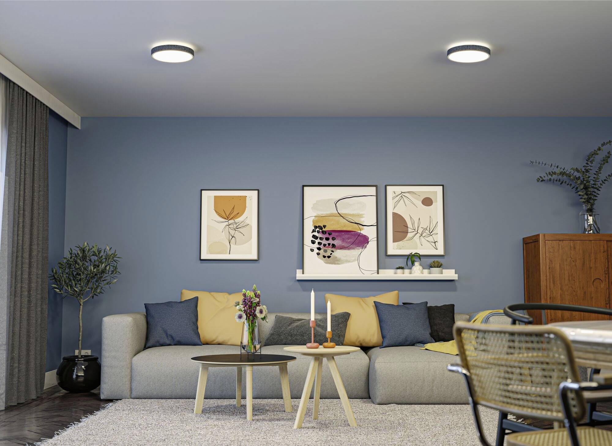 Obývacia izba s modrou stenou, sivou sedačkou, farebnými vankúšmi, dvoma okrúhlymi konferenčnými stolíkmi s dekoráciou a abstraktnou maľbou na stene.