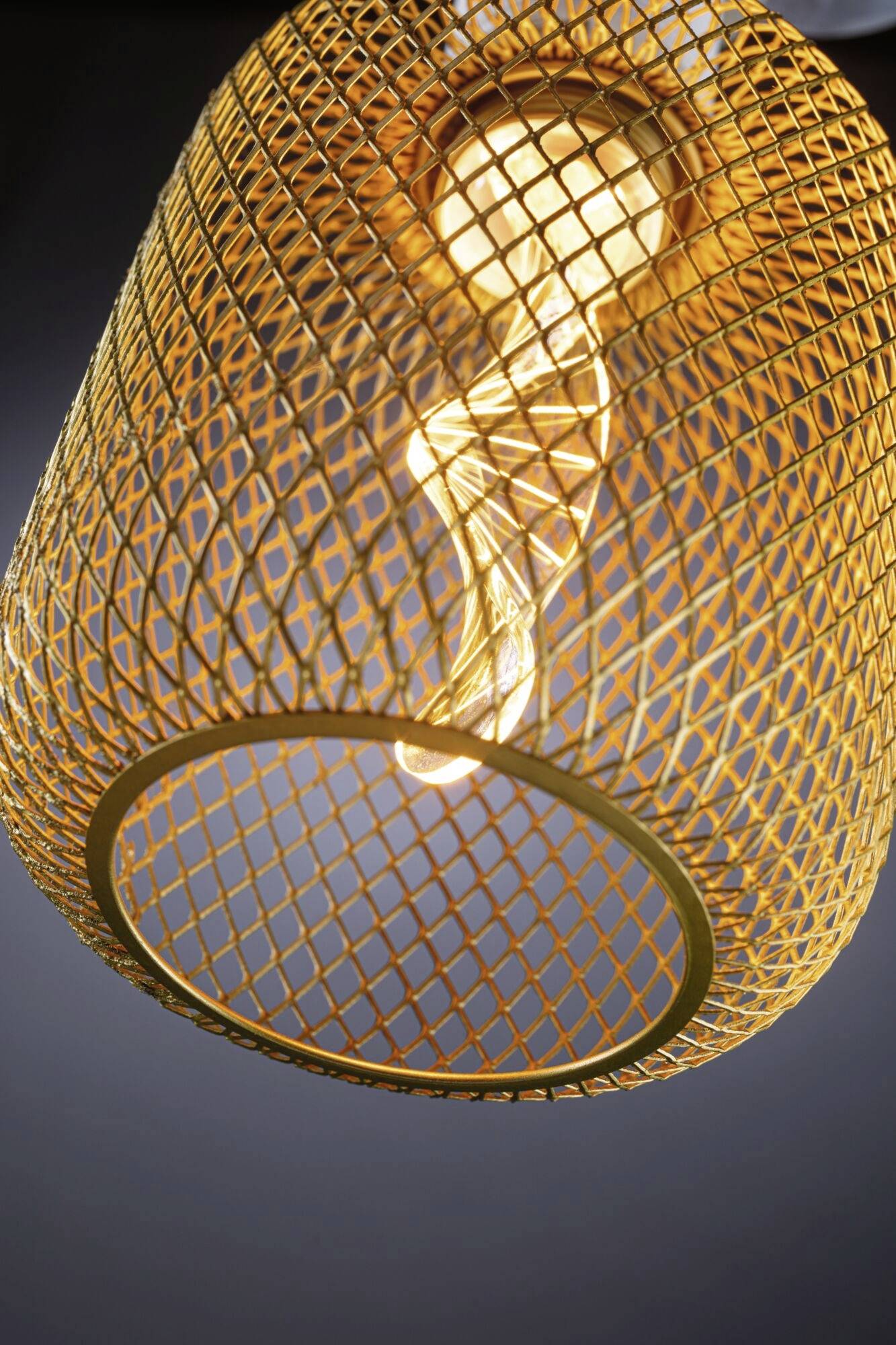 Žltá pletená lampa so žiarovkou, ktorá vyžaruje teplé svetlo, pred tmavým pozadím.