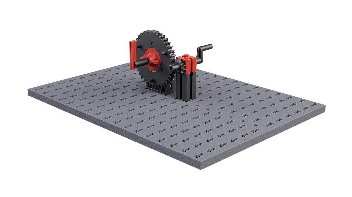 Mechanické ozubené koleso a kľuka na sivej dierovanej doske znázorňujú jednoduchý mechanizmus na prenos sily.