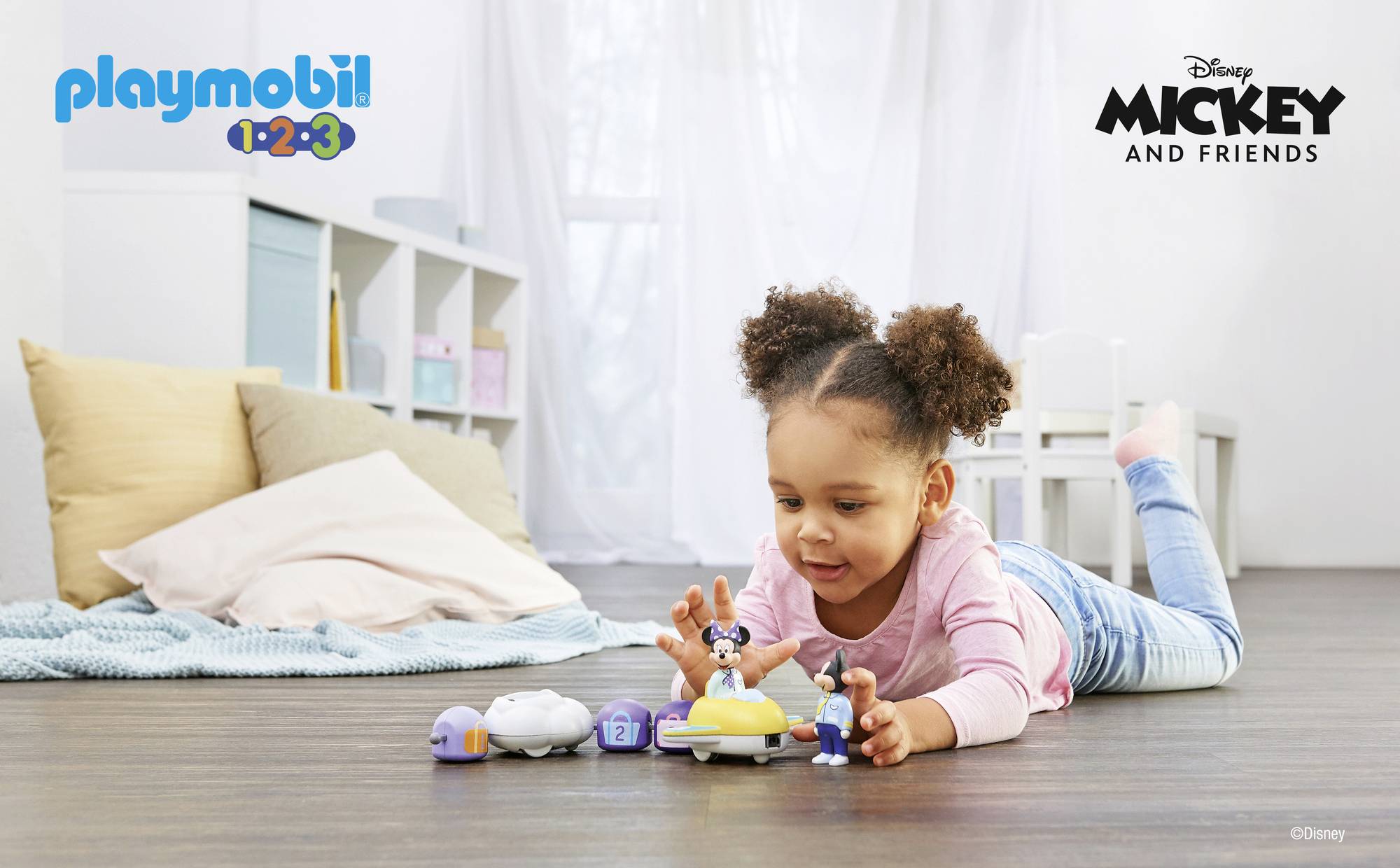 Dieťa sa hrá na drevenej podlahe s hračkami Playmobil z kolekcie 'Disney Mickey and Friends'. V pozadí sú vankúše a police.