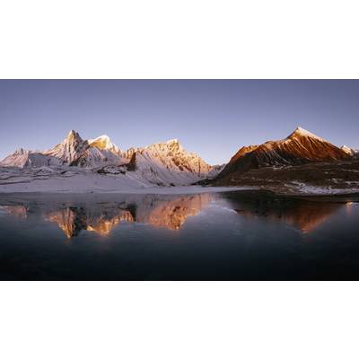 Horská panoráma so zasnežanými vrcholmi a pokojným jazerom v popredí, odrážajúca teplé svetlo zapadajúceho slnka.