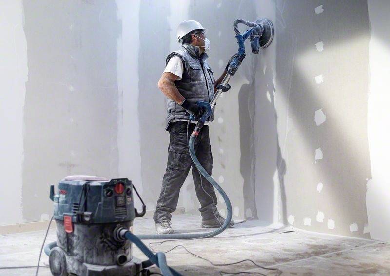 Robotník v ochrannej prilbe a maske vyhladzuje stenu brúskou v miestnosti. Vedľa neho stojí vysávač.