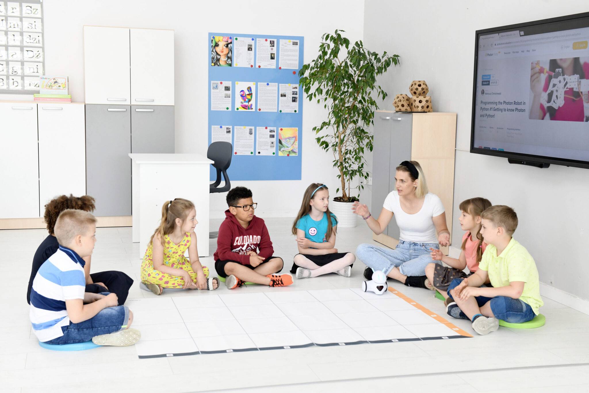 Učiteľka sedí v polkruhu s deťmi v triede. Na obrazovke sú pedagogické obsahy. V pozadí je nástenka.