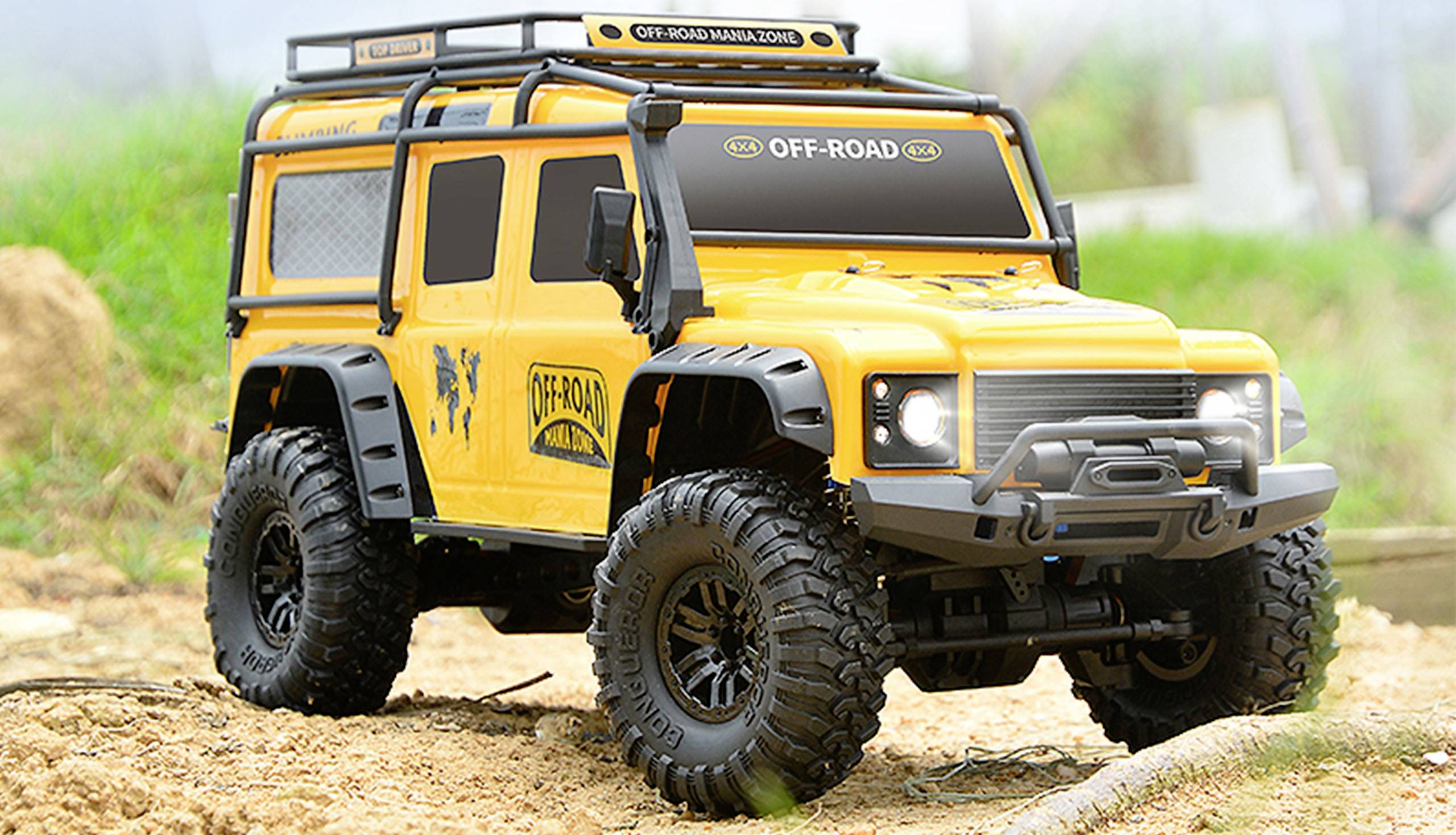 Žlté diaľkovo ovládané terénne vozidlo s nápisom 'OFF-ROAD' stojí na piesknatom teréne, obklopené trávou a stromami v pozadí.