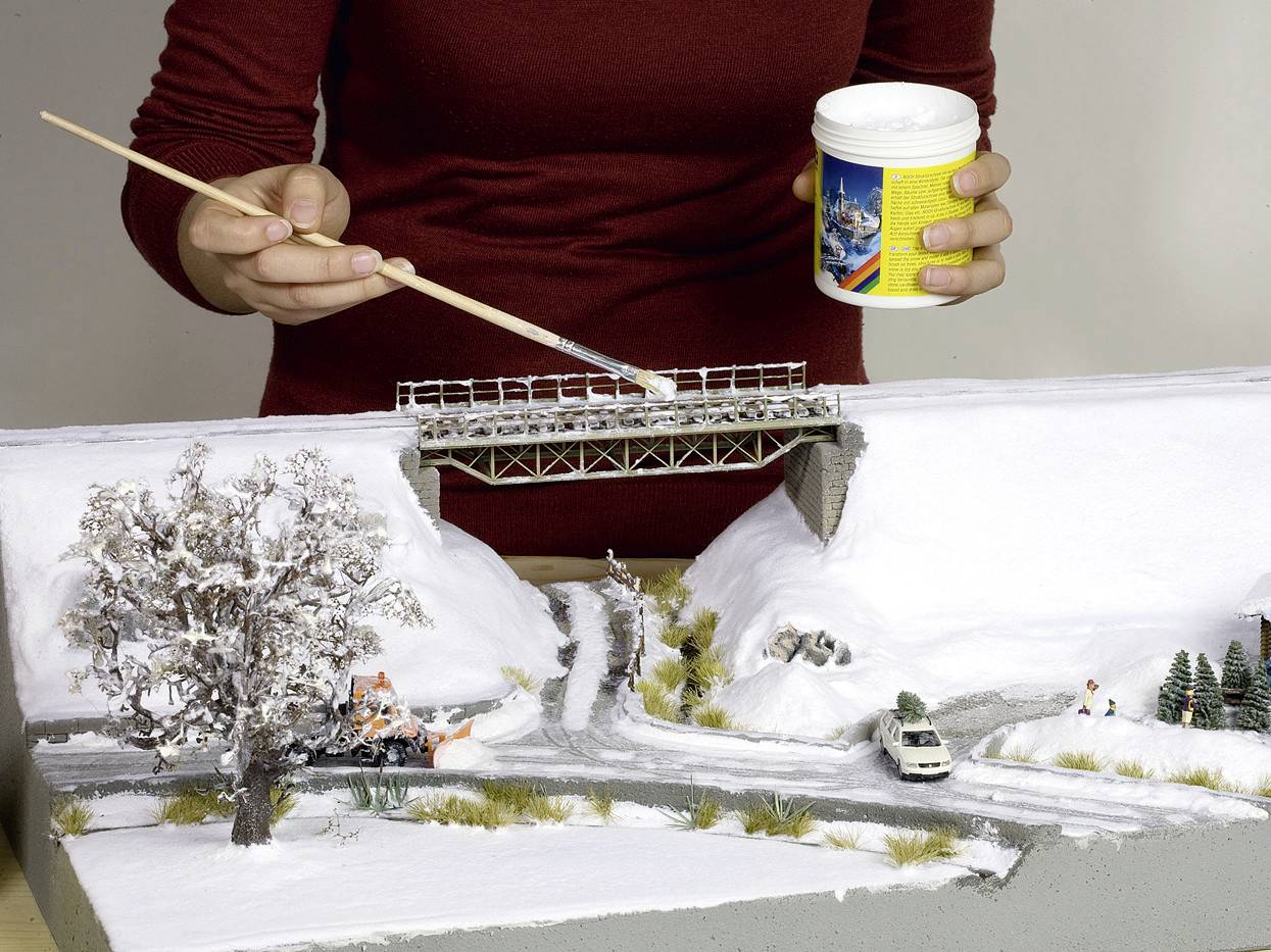 Ruka maľuje štetcom sneh na miniatúrny model zimnej krajiny s mostom, cestou, stromami a autami.