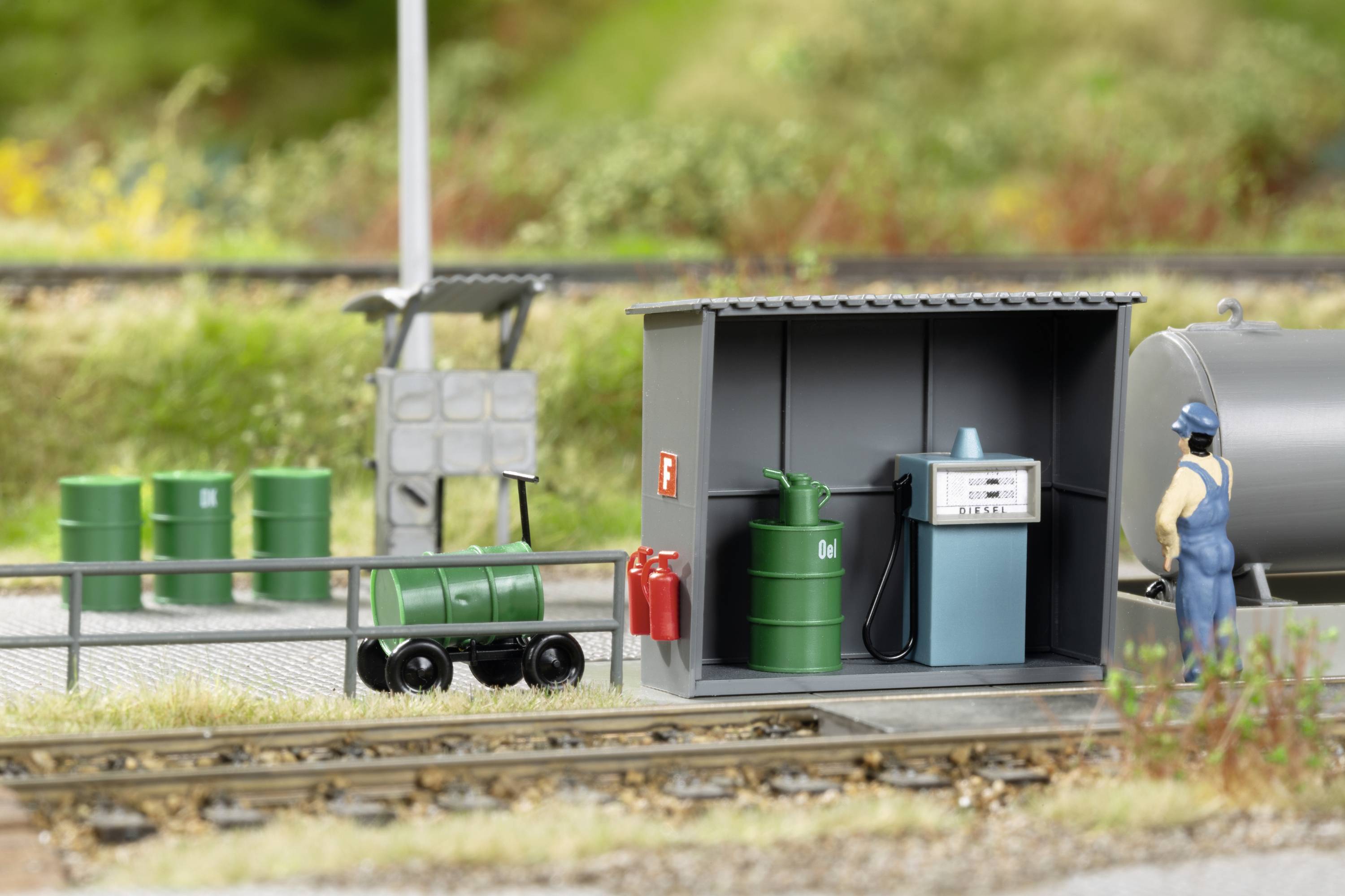 Model železničnej čerpacej stanice so zeleným cisternovým vozňom, čerpacou stanicou a zamestnancami v modrom oblečení, obklopený železničnými koľajami.
