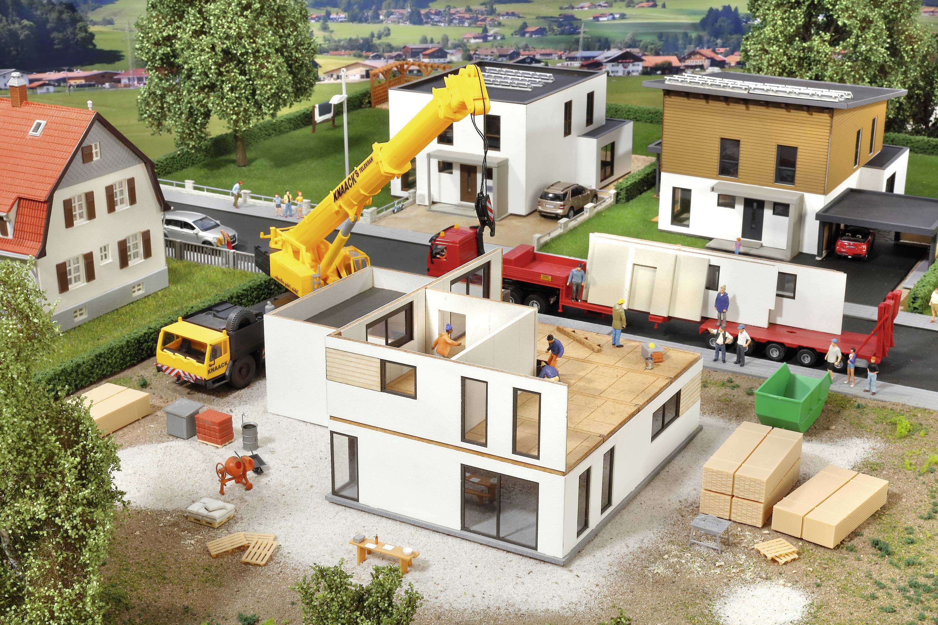 Model staveniska zobrazuje výstavbu domu s žeriavom, nákladným autom a robotníkmi, obklopený niekoľkými dokončenými domami.