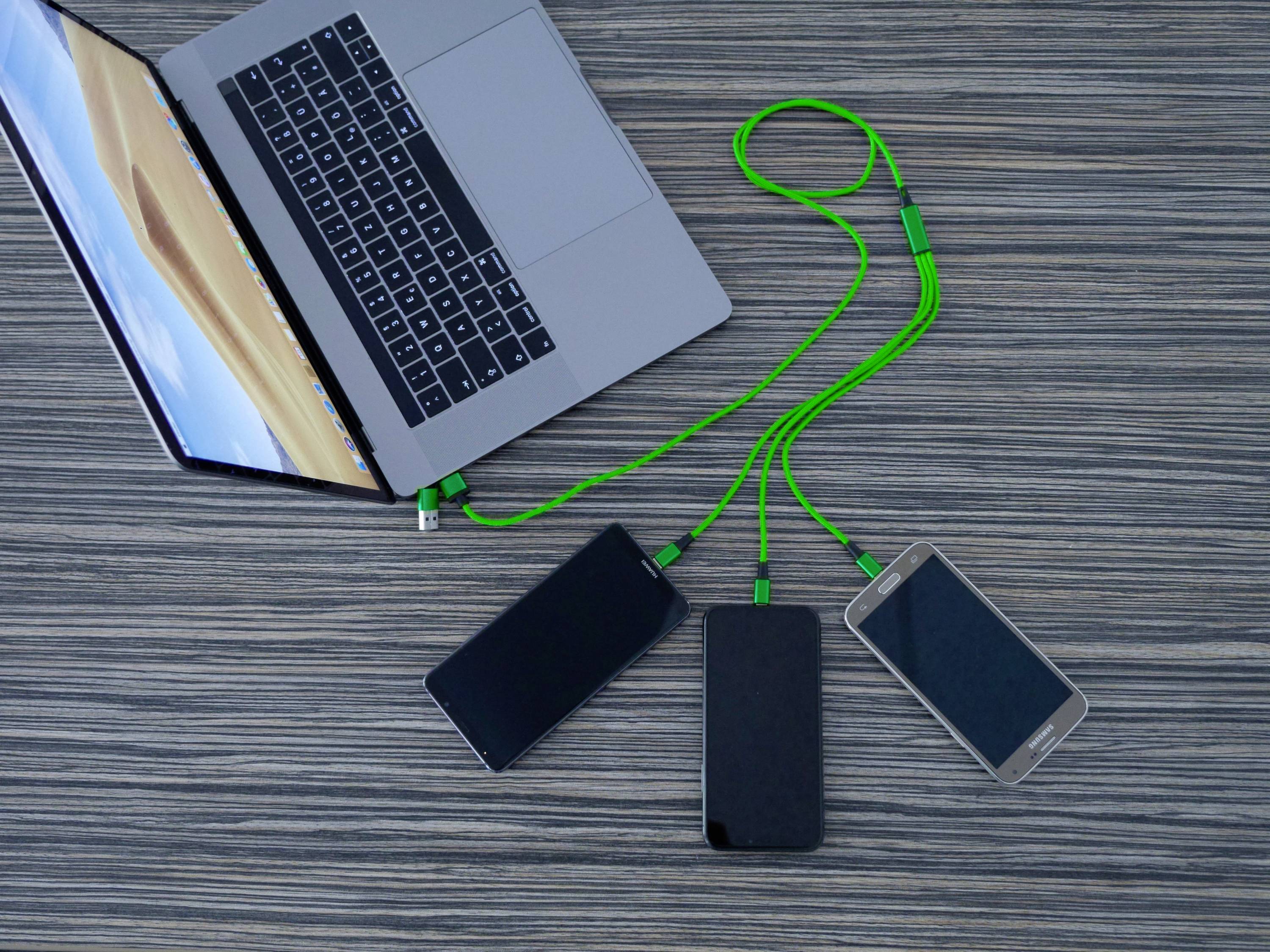 Laptop s zeleným káblom, ktorý nabíja tri smartfóny. Všetky zariadenia ležia na drevenom stole.