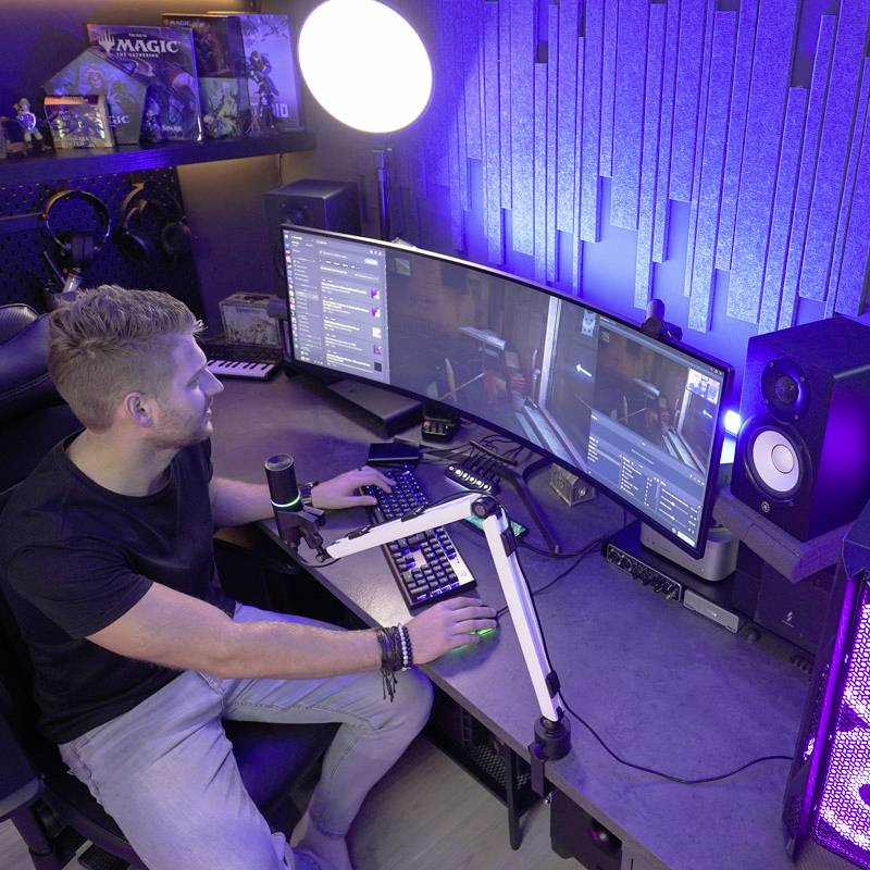 Muž sedí pri stole s viacerými monitormi a mikrofónom, pričom hrá počítačové hry alebo streamuje.