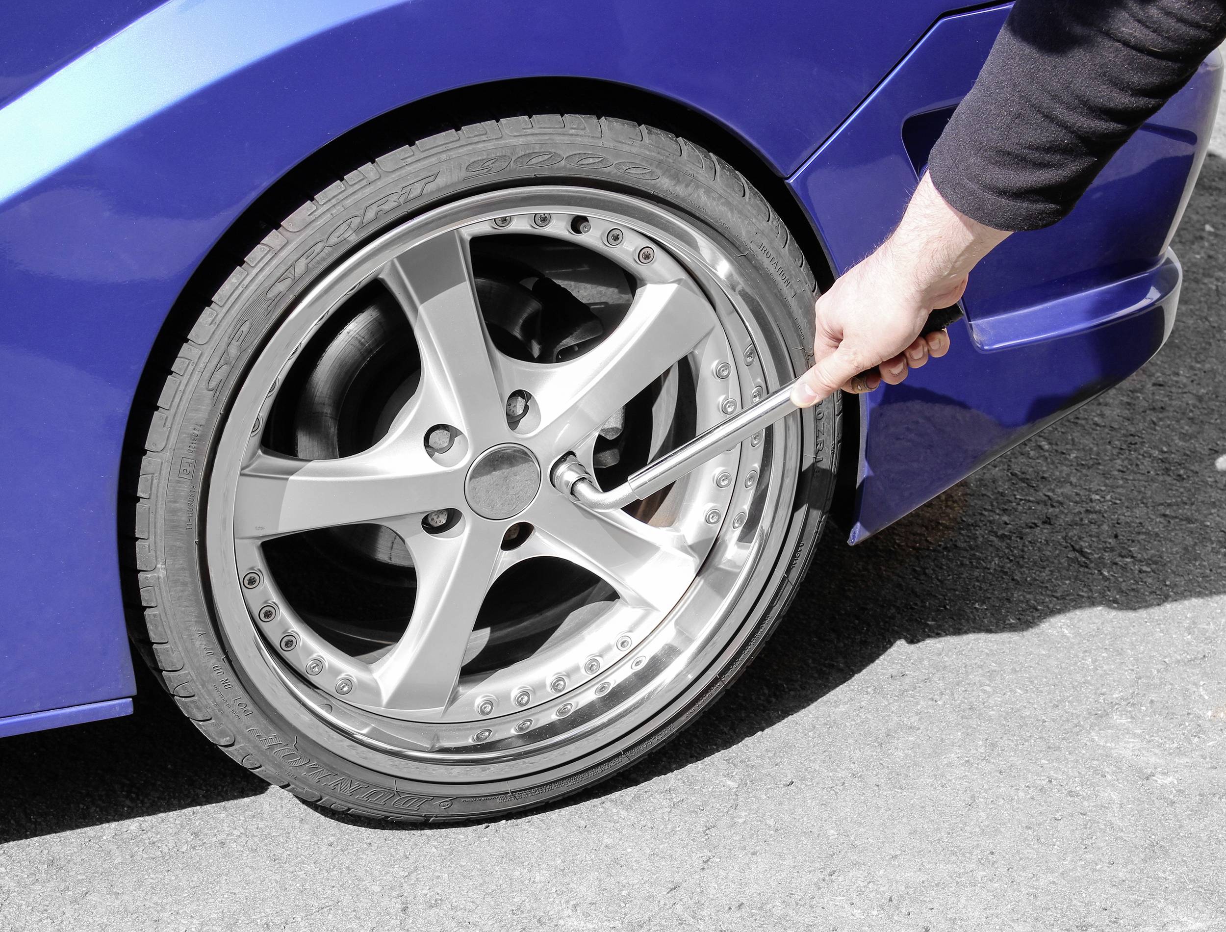 Osoba uťahuje maticu kolesa kľúčom na modrom aute. Auto stojí na asfaltovej ploche.