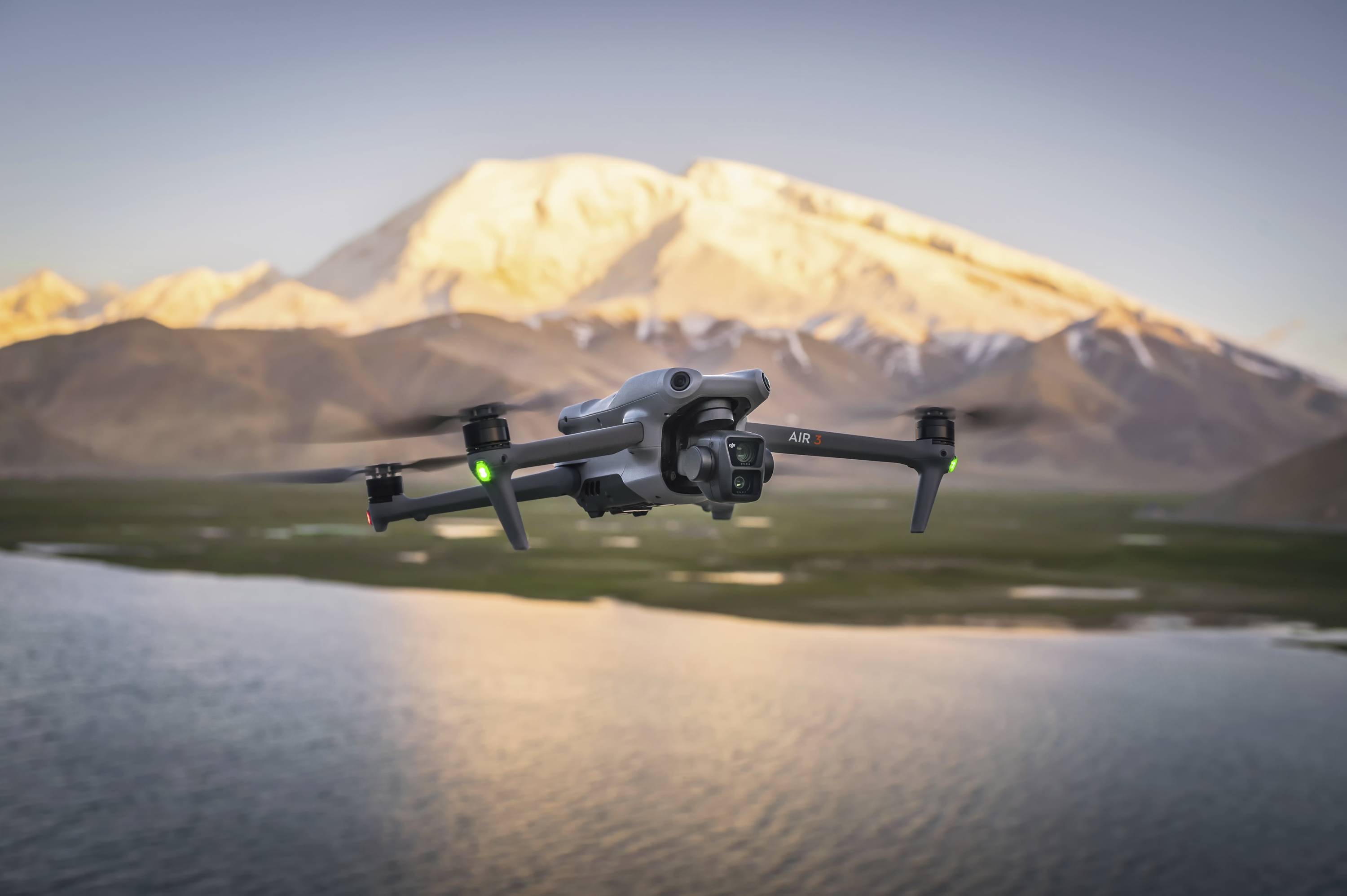 Dron letí nad jazerom, v pozadí sú viditeľné zasnežené hory. Dron má zelené a červené svetlá.