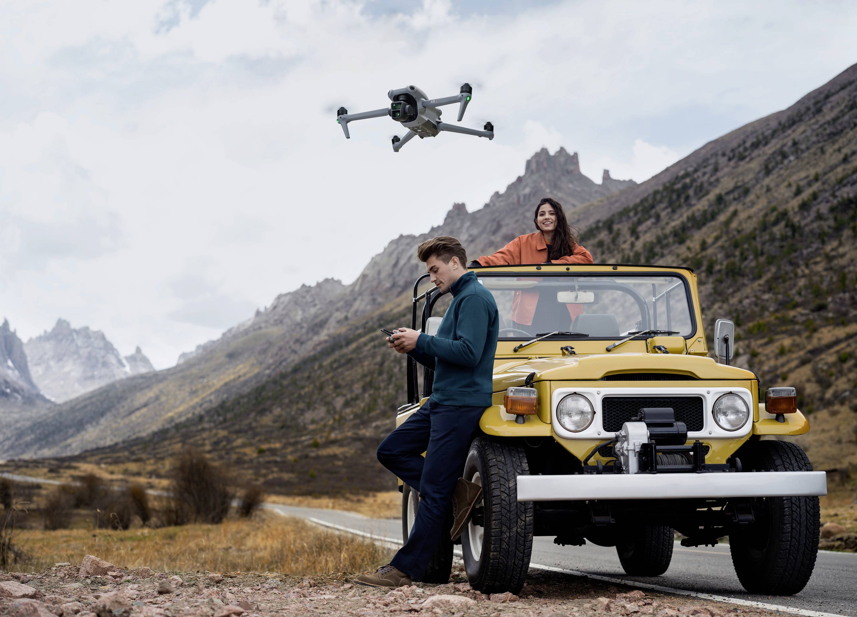 Muž ovláda dron vedľa žltého terénneho vozidla v kopcovitej krajine, pričom žena stojí v aute a usmieva sa.