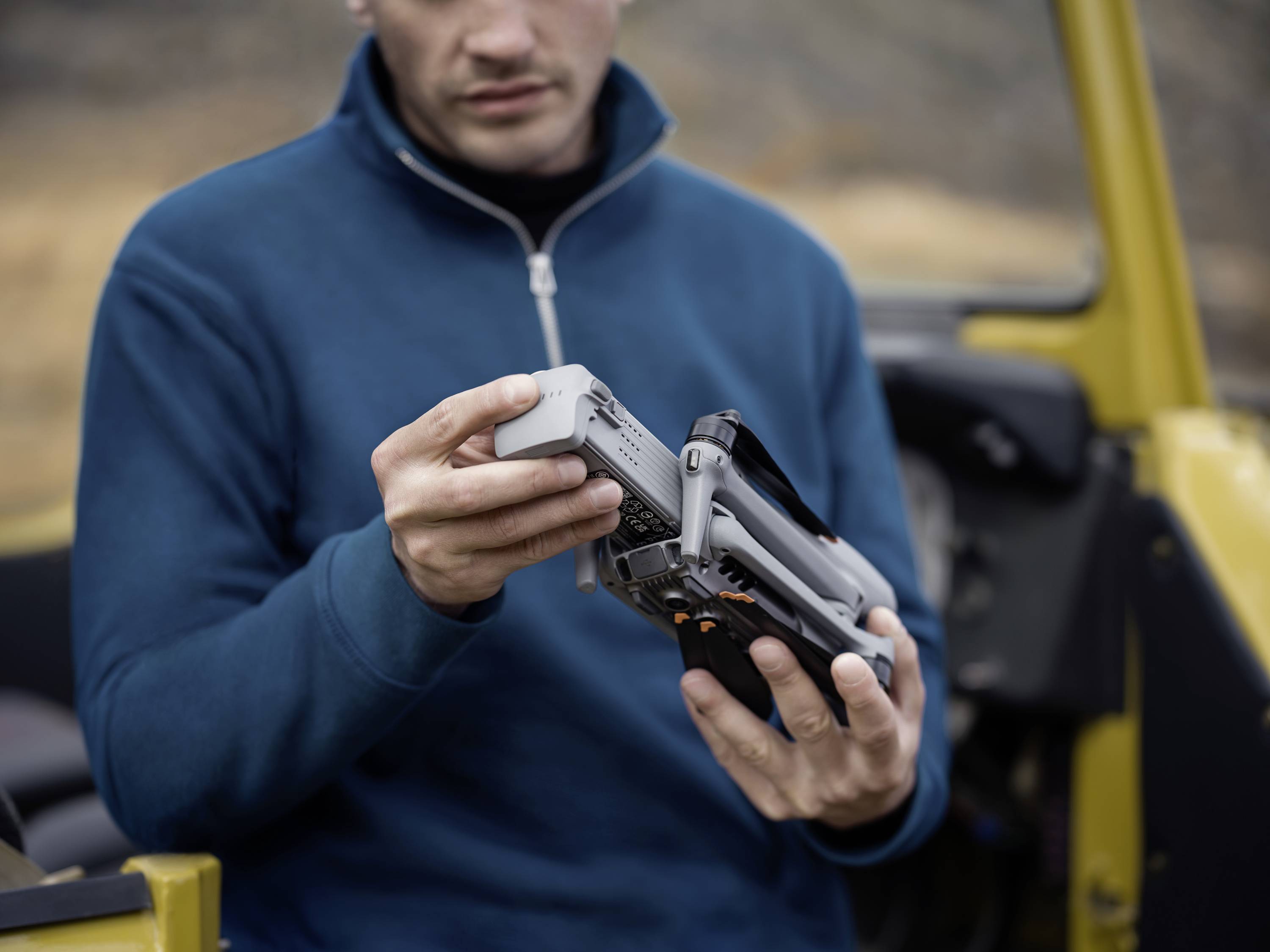 Osoba v modrej bunde drží a kontroluje sivý dron, pravdepodobne pred nasadením v exteriéri.
