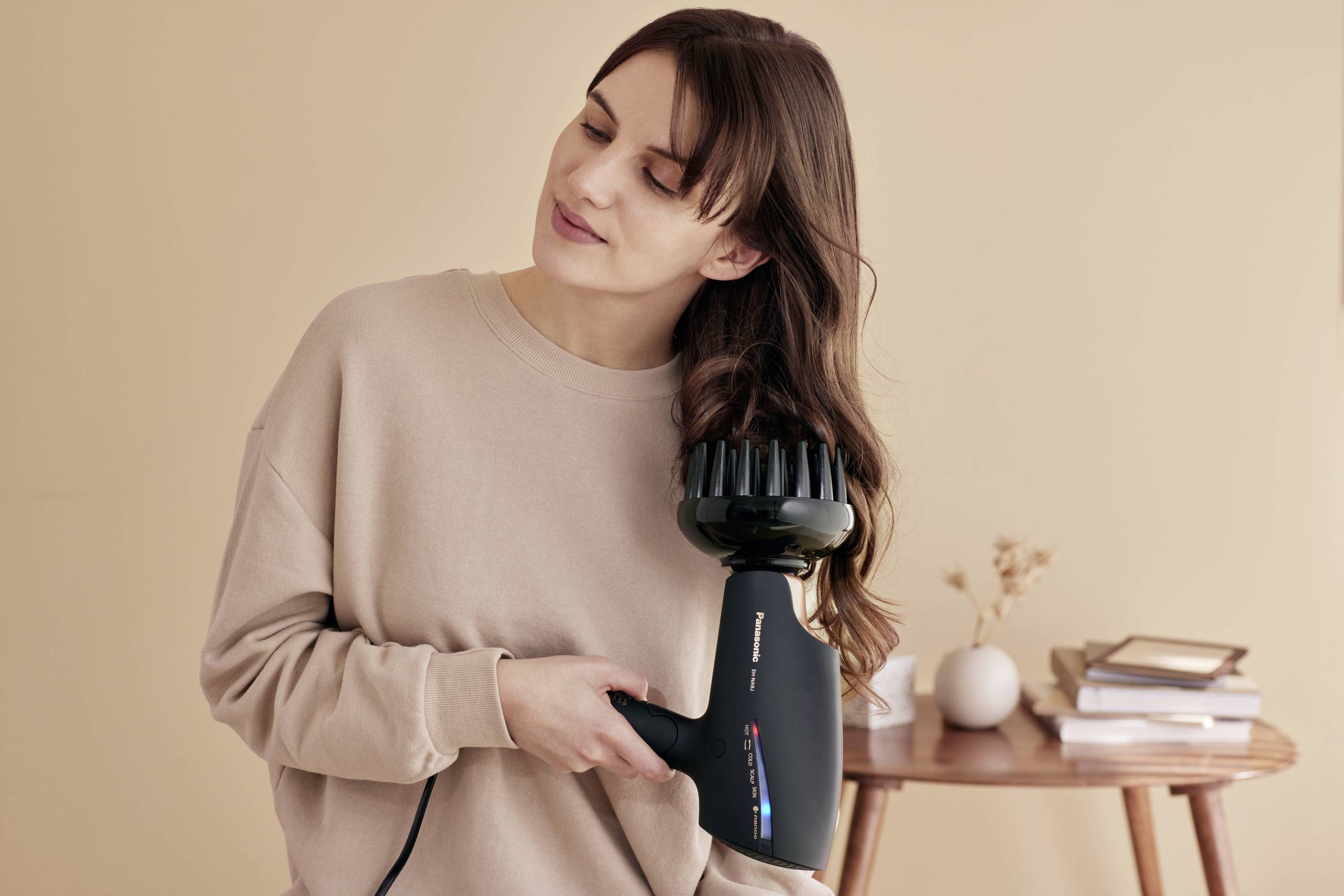Žena s hnedými vlasmi používa čierny fén s difúzorom v miestnosti zariadené v béžových tónoch.