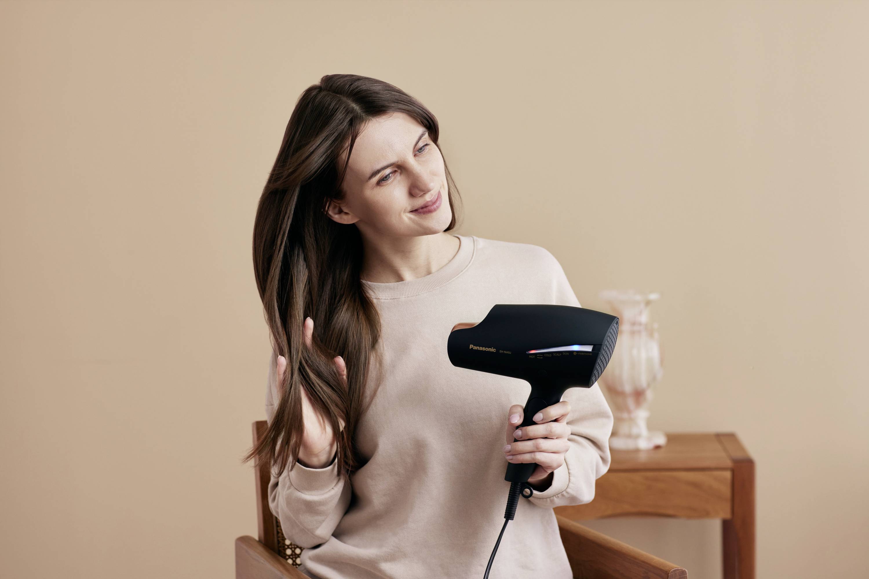 Žena s dlhými hnedými vlasmi suší svoje vlasy. Drží v ruke čierny fén a sedí na stoličke.
