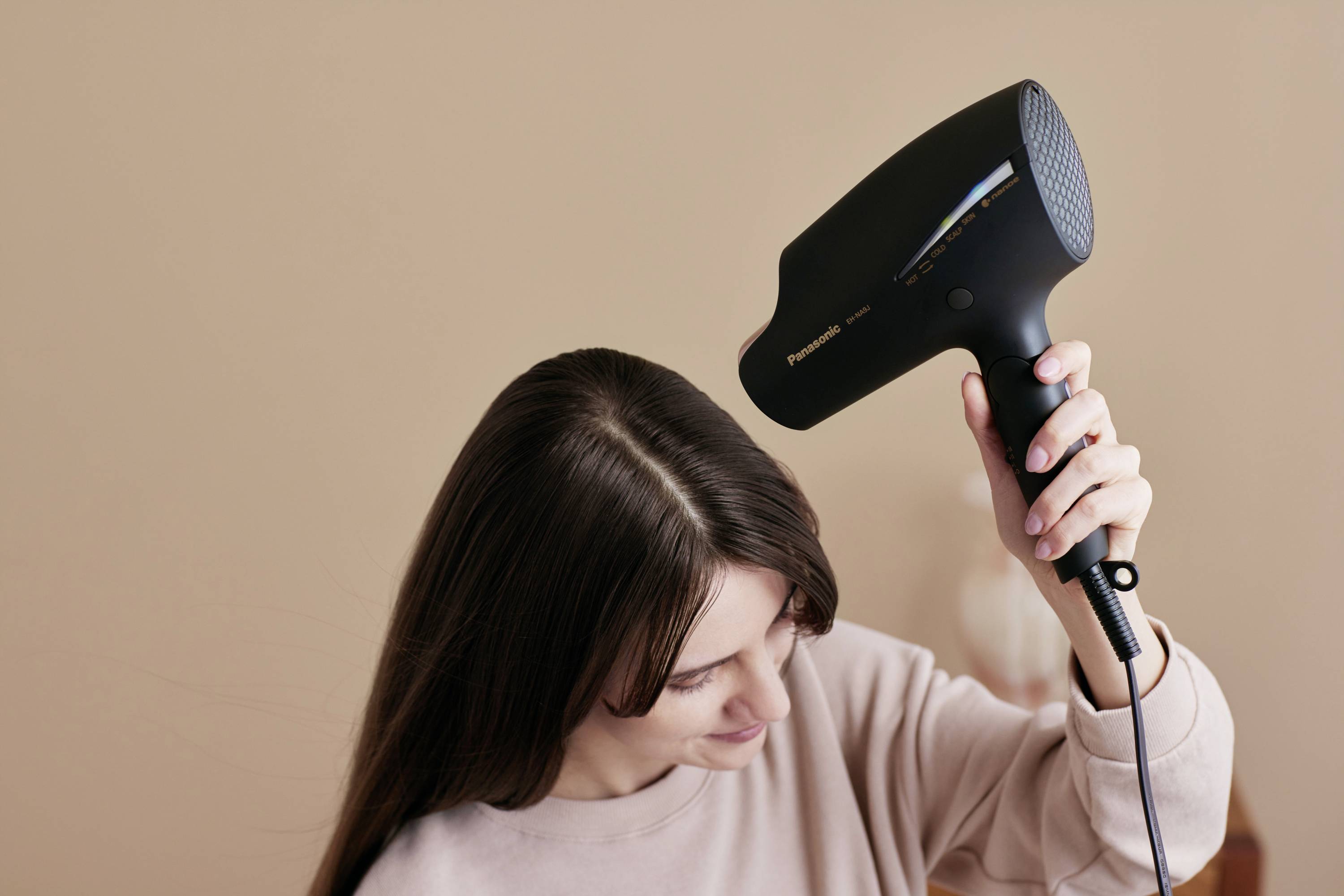 Žena si fénom suší dlhé tmavé vlasy pomocou čierneho sušiča vlasov.
