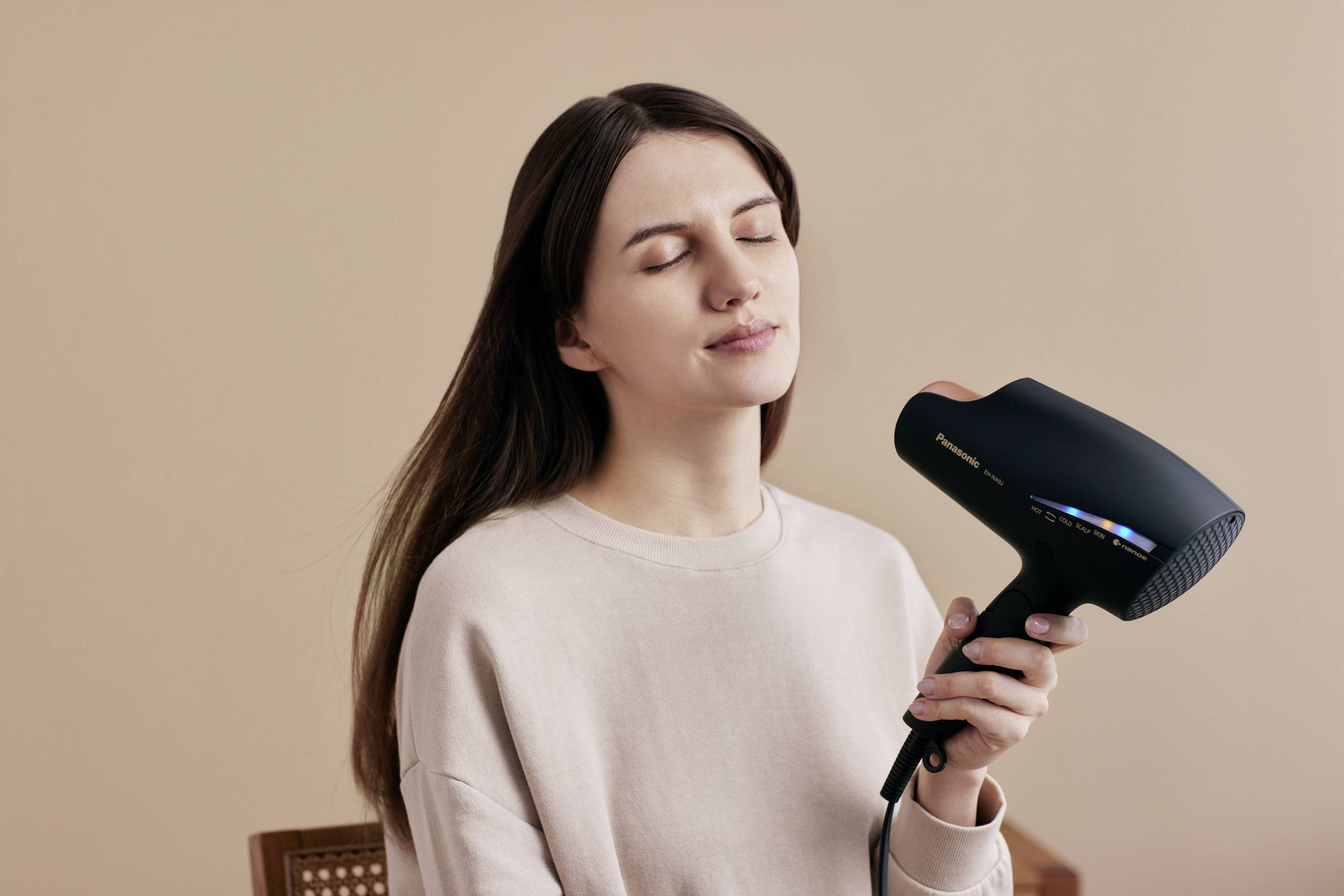 Žena so zatvorenými očami si fénom suší dlhé hnedé vlasy pred béžovým pozadím.