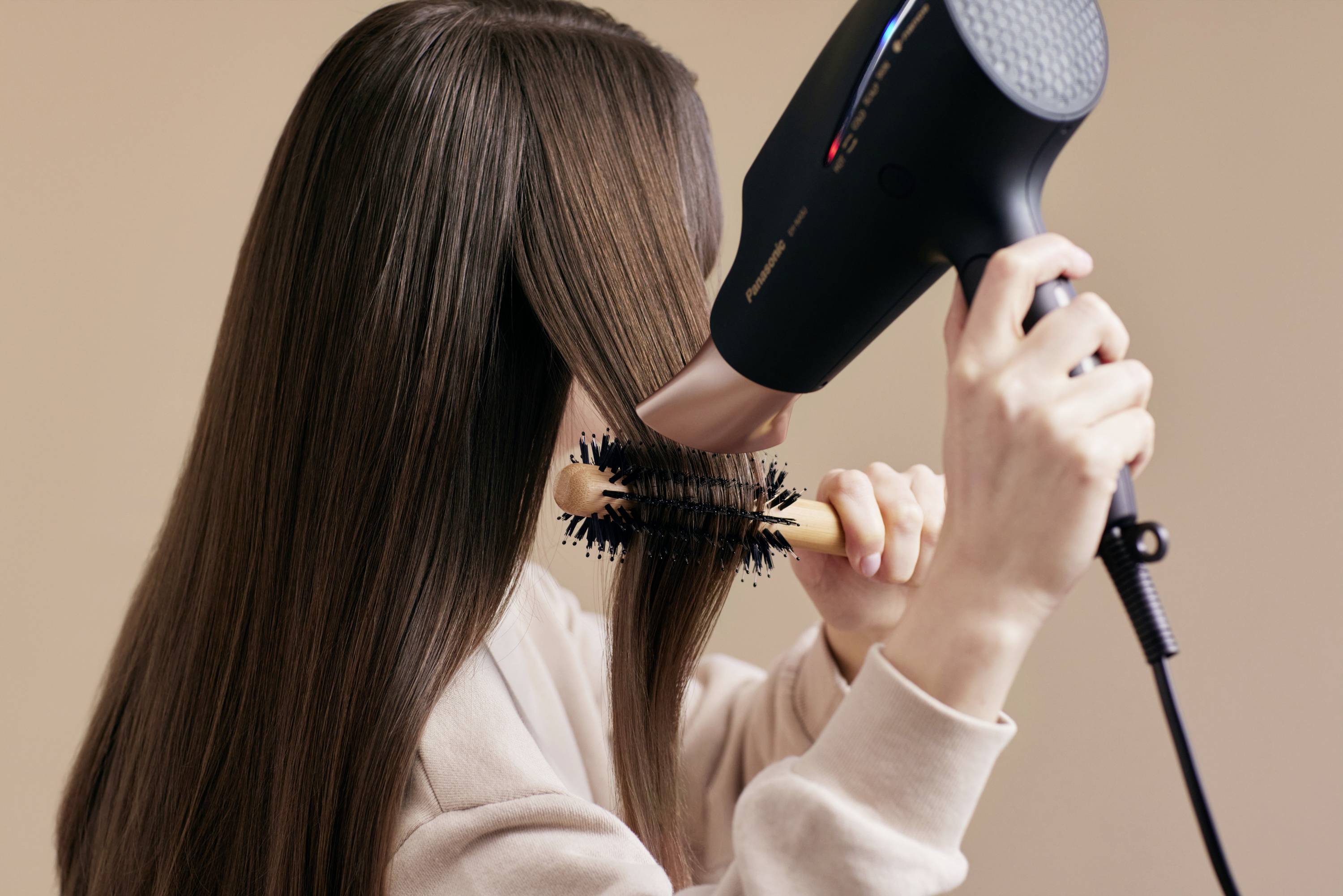 Žena si fénom suší dlhé, hladké vlasy a zároveň ich tvaruje okrúhlou kefou.