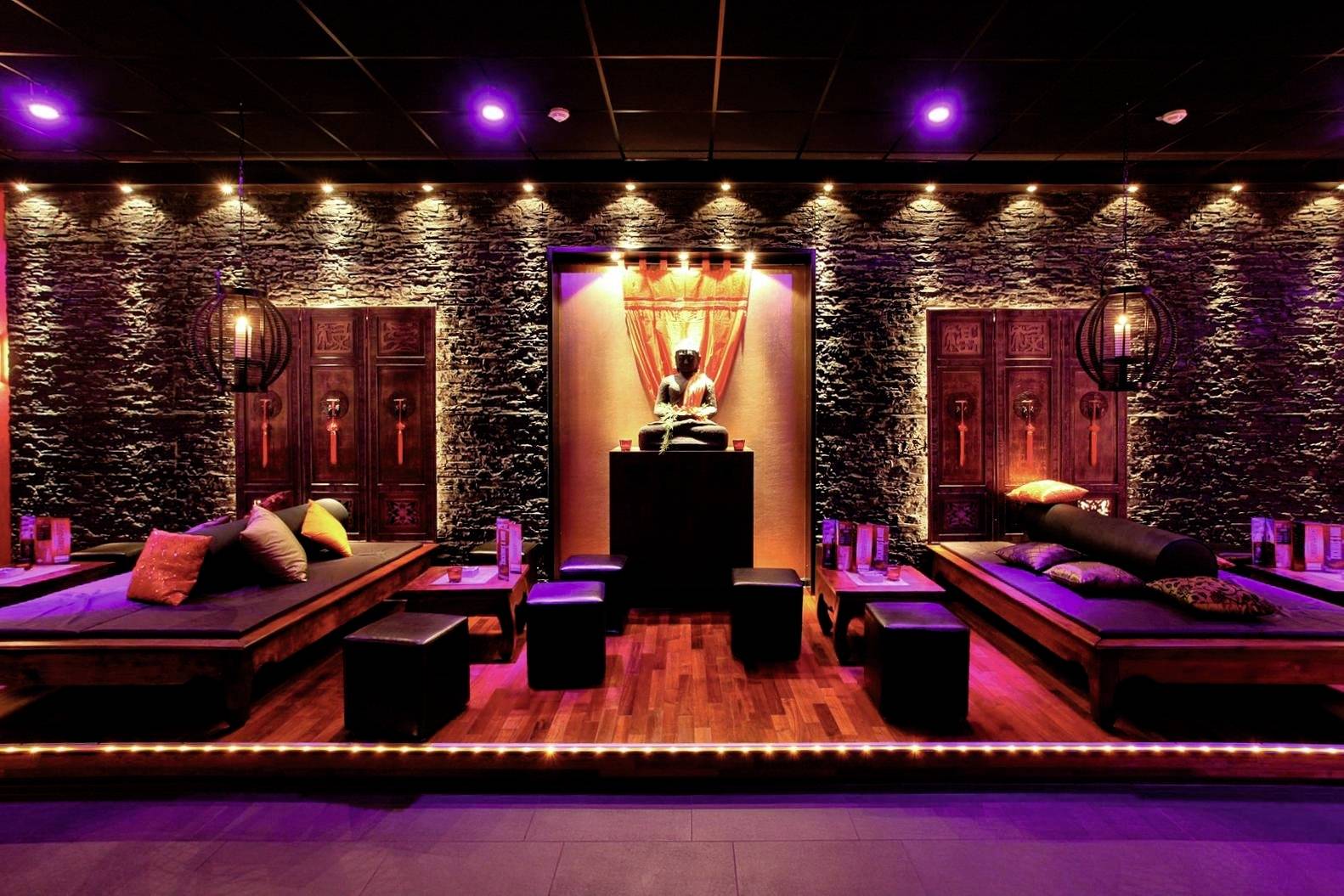 Štýlovo osvetlený oddychový priestor s nízkymi sedadlami a dekoratívnymi vankúšmi, obklopený kamennými stenami a teplým svetlom.