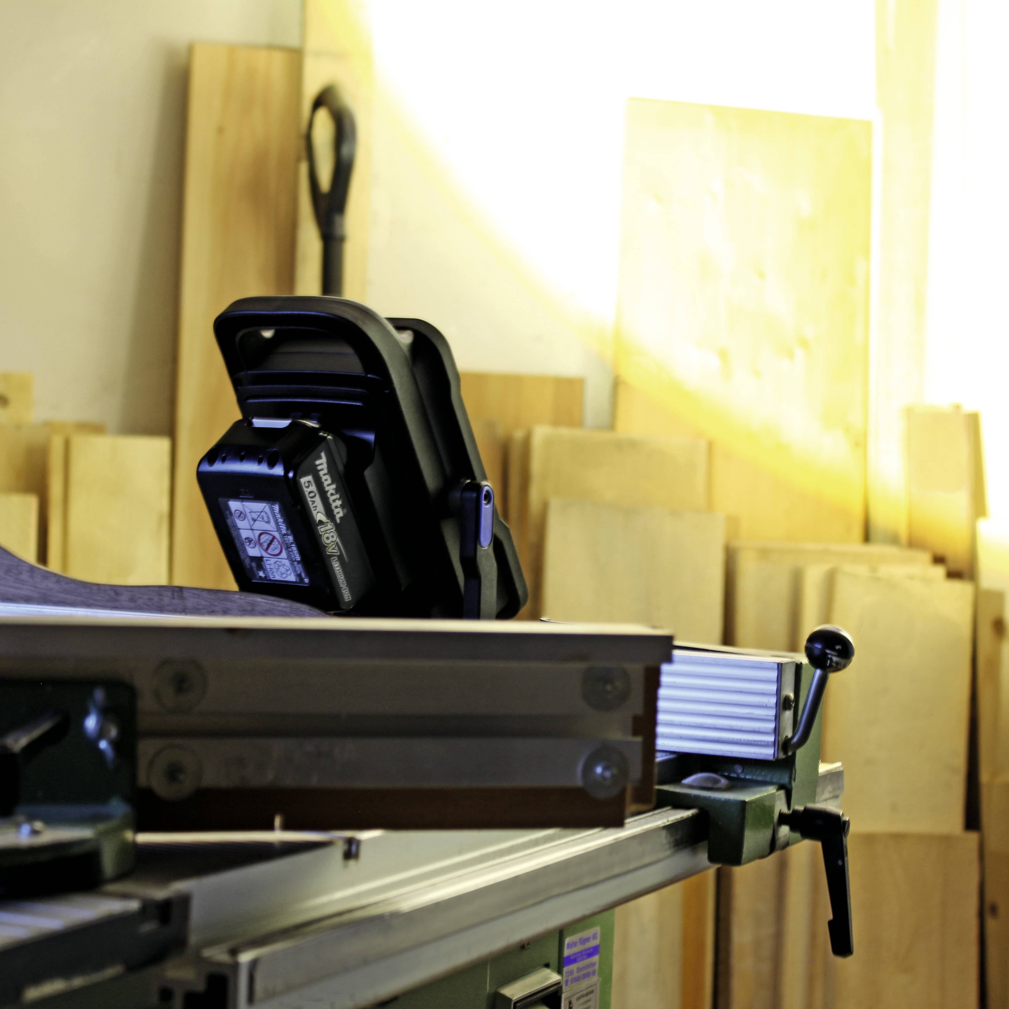 V dielni stojí stolná kotúčová píla. V pozadí sú viditeľné drevené dosky a náradie. Na píle leží meter.