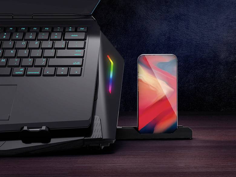 Laptop s podsvietením klávesnice je pripojený k smartfónu, ktorý stojí v držiaku. Obe zariadenia sú umiestnené na drevenom stole.