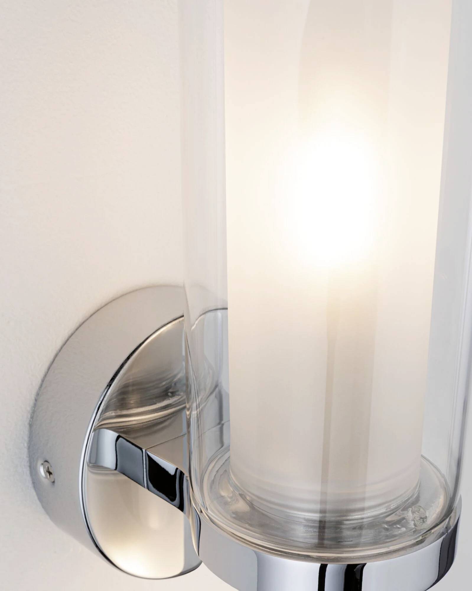 Svietiaca nástenná lampa s moderným dizajnom, valcovitým skleneným tienidlom a strieborným podstavcom, namontovaná na svetlej stene.