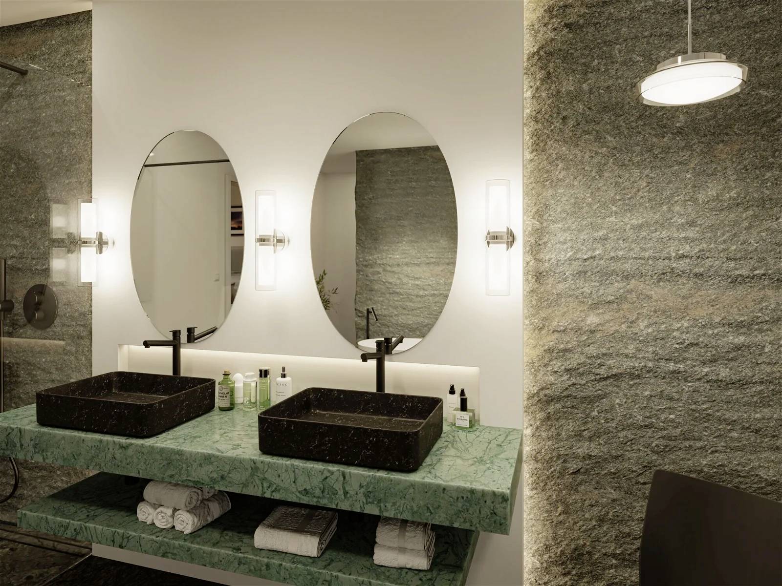 Moderná kúpeľňa s dvoma oválnymi zrkadlami nad zelenou mramorovou doskou s dvoma čiernymi umývadlami a kozmetickými produktmi.