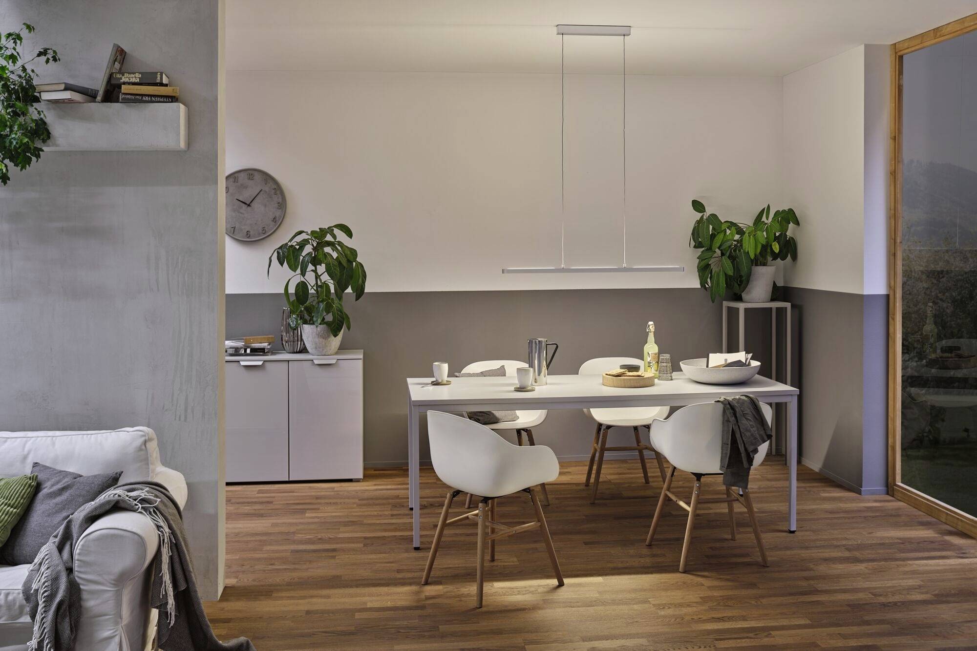 Jedáleň s bielym stolom, štyrmi stoličkami a modernou dekoráciou. Rastliny po stranách, hodiny na stene, minimalistické osvetlenie.