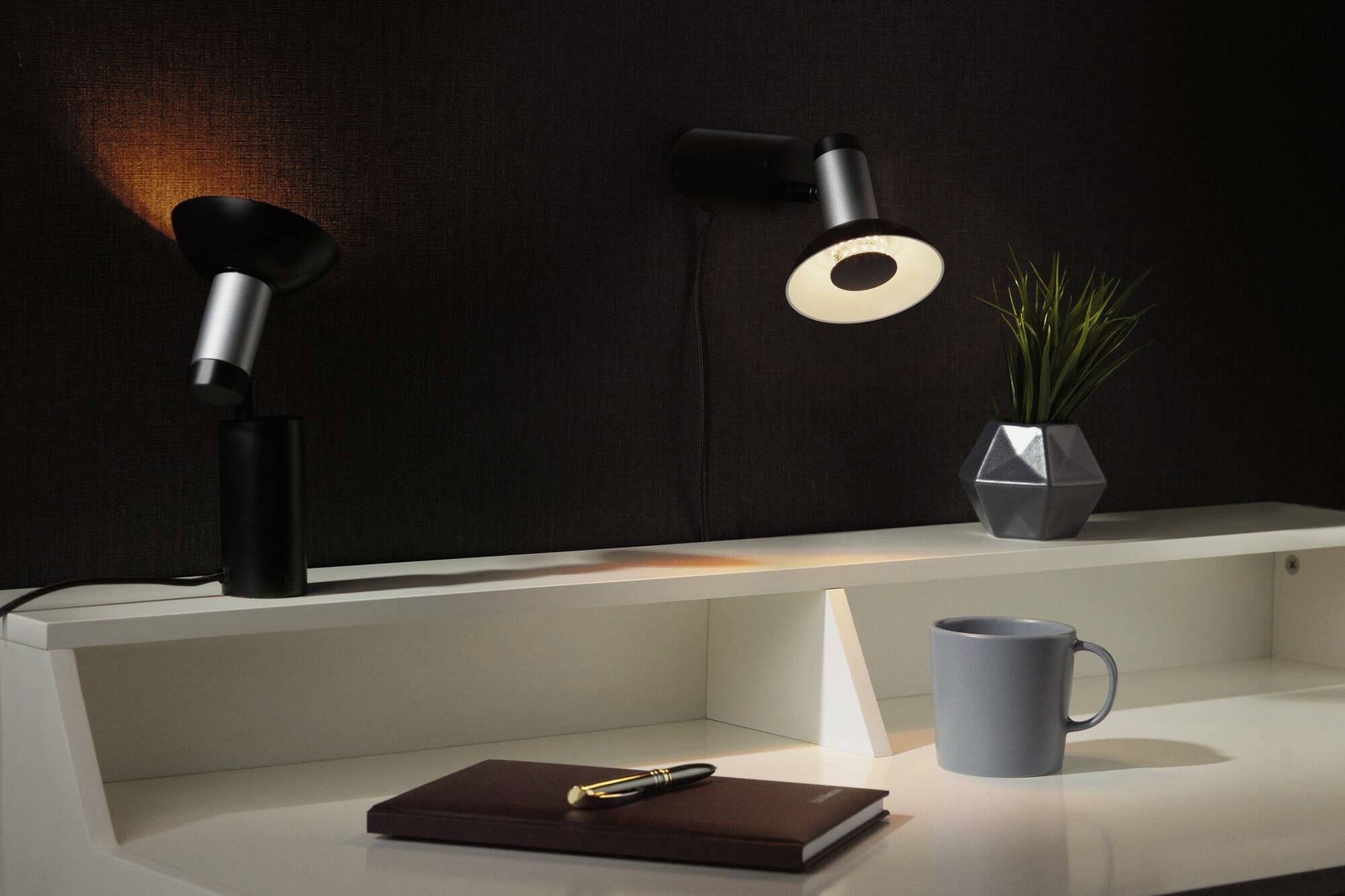 Dve moderné stolové lampy na bielom písacom stole osvetľujú poznámkový zošit s perom, šálku kávy a rastlinu v geometrickej nádobe.