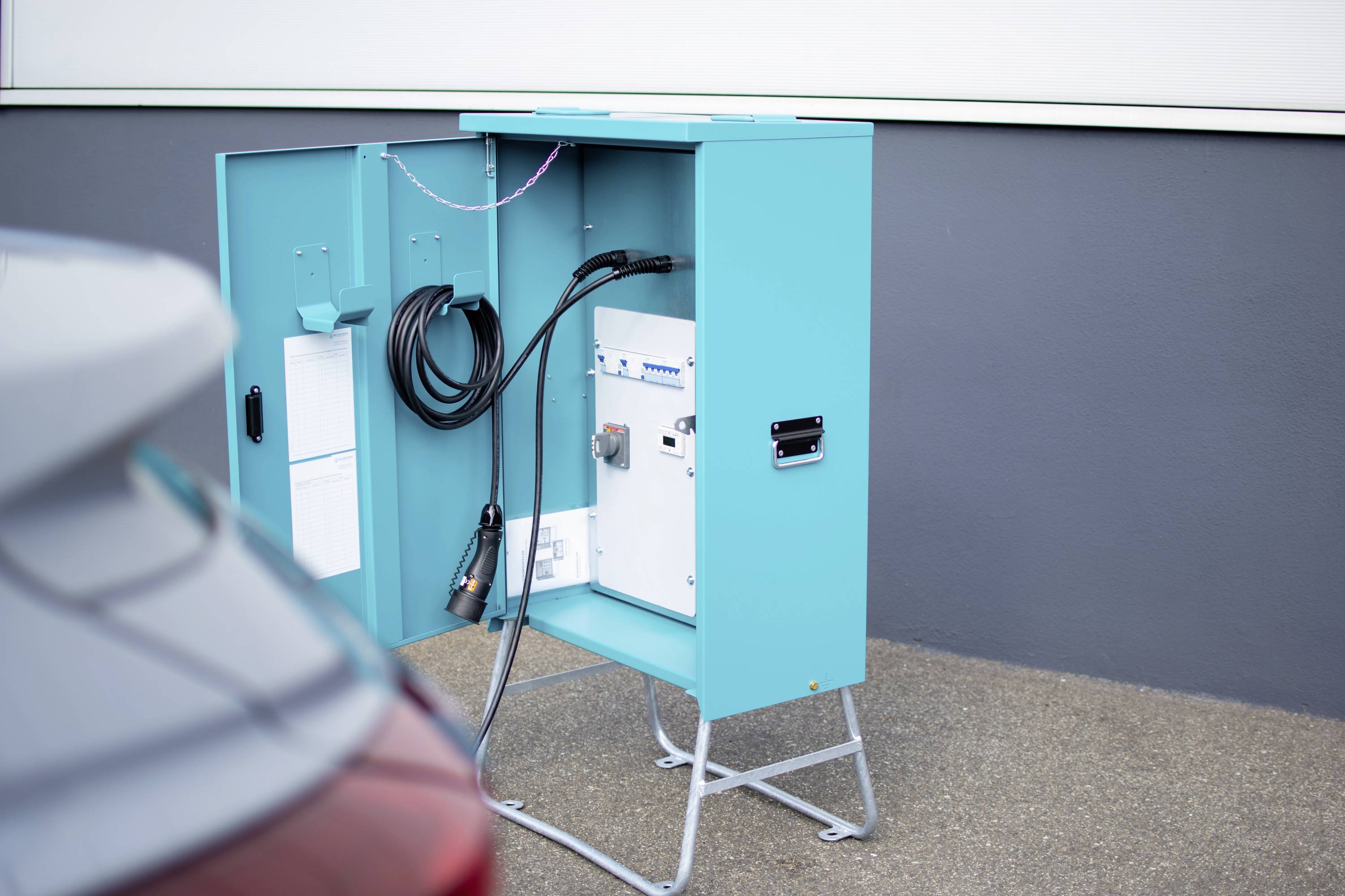 Otvorená tyrkysová krabica s káblami a elektronikou, pravdepodobne nabíjacia stanica, stojí na sivej podlahe vedľa vozidla.