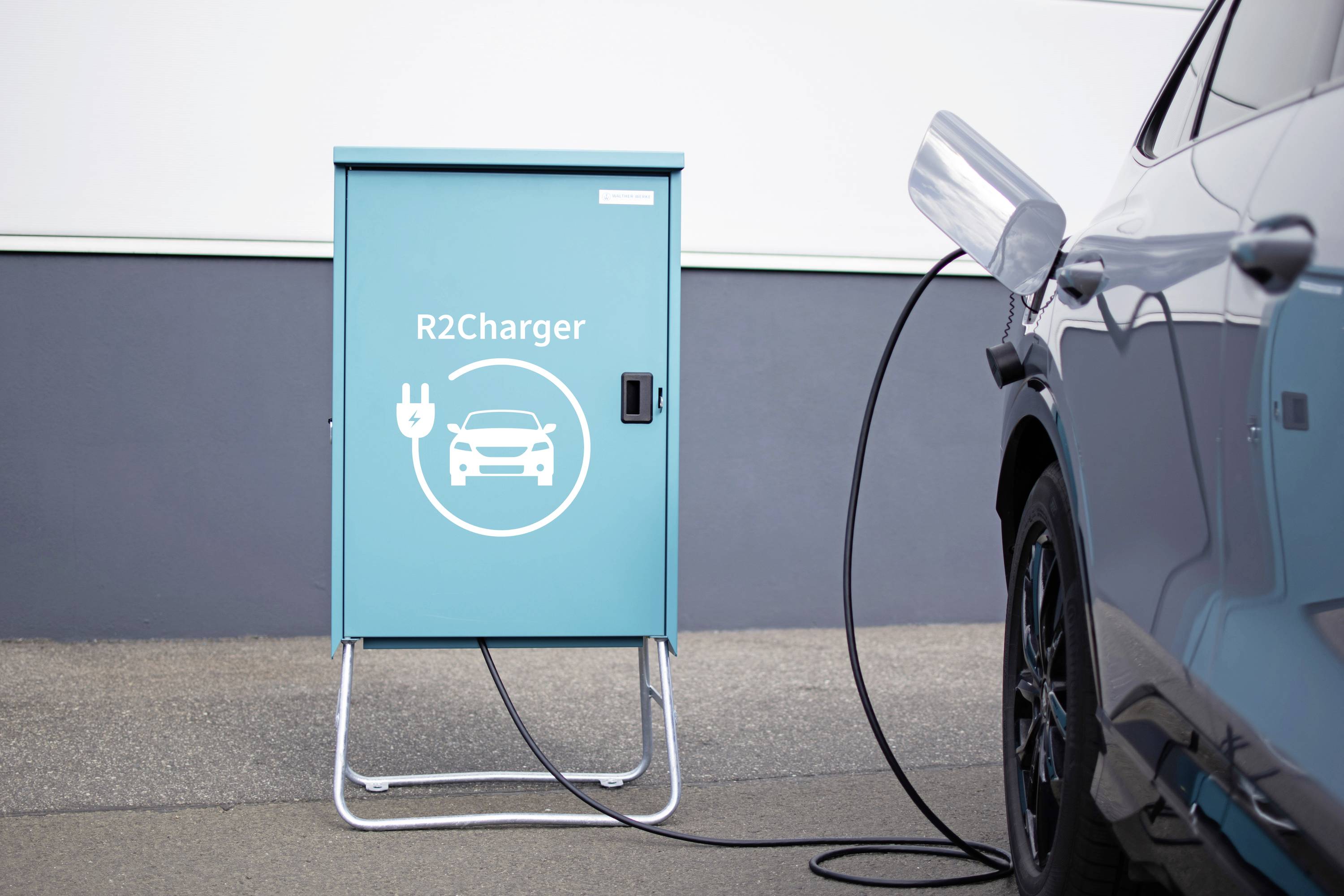 Elektrické auto je pripojené k nabíjacej stanici s názvom 'R2Charger' prostredníctvom nabíjacieho kábla. Lokalita je mestská.