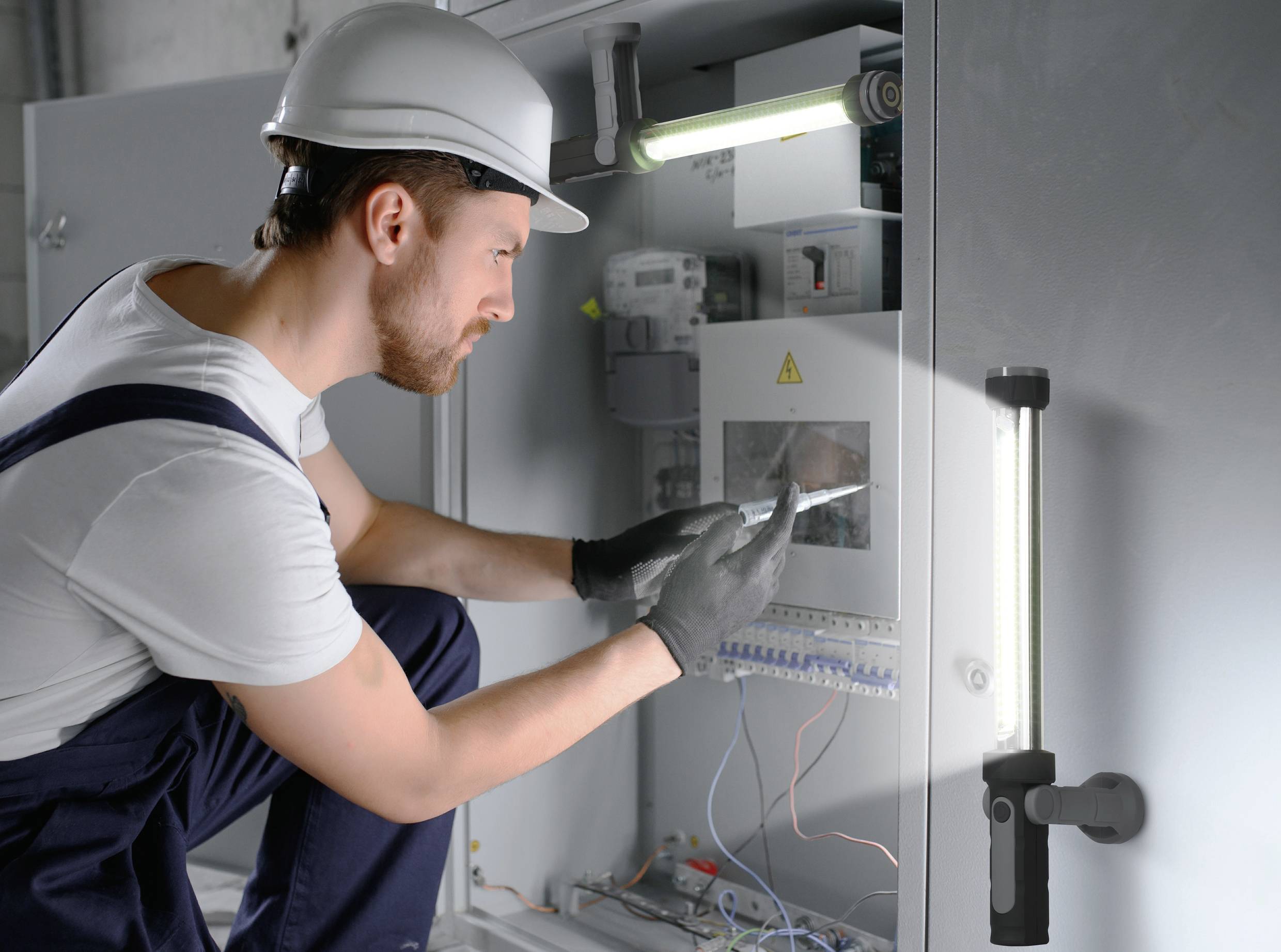 Elektrikár v pracovnom oblečení a s helmou kontroluje rozvádzač pomocou tabletu, obklopený káblami a náradím.
