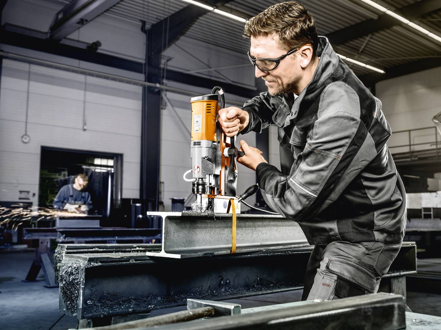 Muž v pracovnom oblečení obsluhuje magnetickú vŕtačku na oceľovom nosníku v dielni. V pozadí pracuje ďalšia osoba.