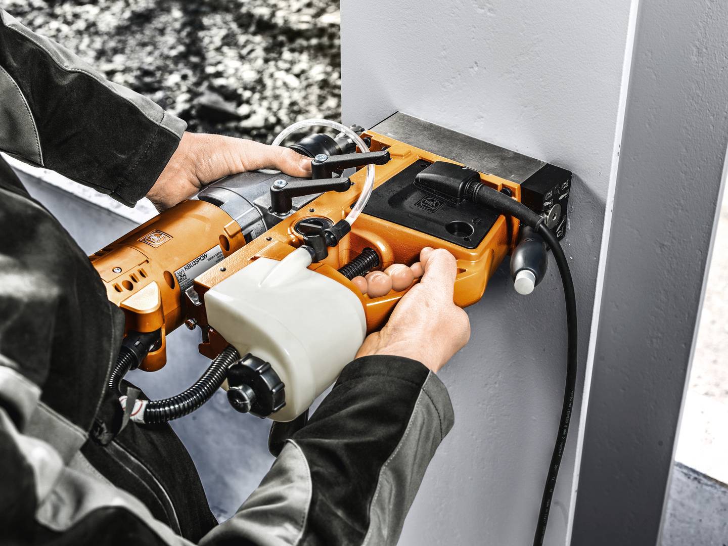 Osoba používa oranžovú vŕtačku na vŕtanie do šedého kovového nosníka. V rozmazanom pozadí je viditeľná stavenisko.