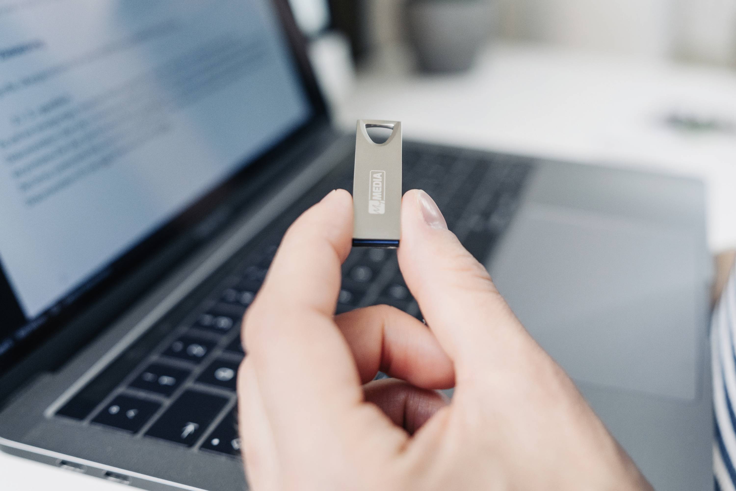 Osoba drží USB kľúč pred otvoreným laptopom. Na obrazovke je rozmazaný textový dokument.