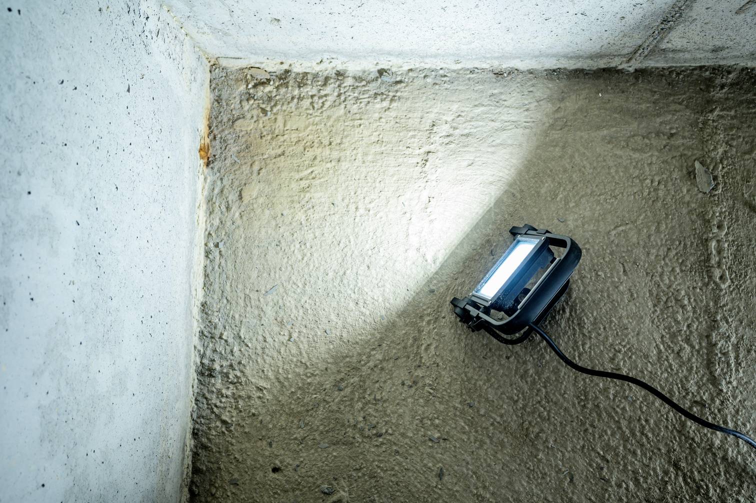Prenosná lampa osvetľuje kút s drsnou betónovou podlahou a stenami, ktoré reprezentujú industriálny alebo nedokončený priestor.