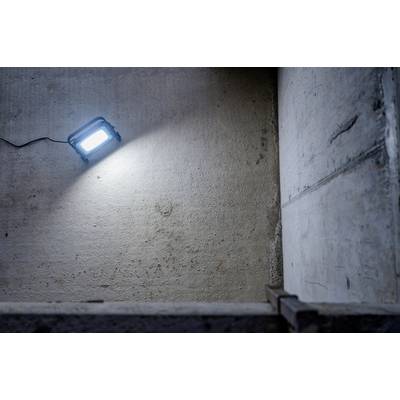 Modrý stavebný reflektor osvetľuje sivú betónovú stenu v prázdnej miestnosti. Kábel vedie doľava.