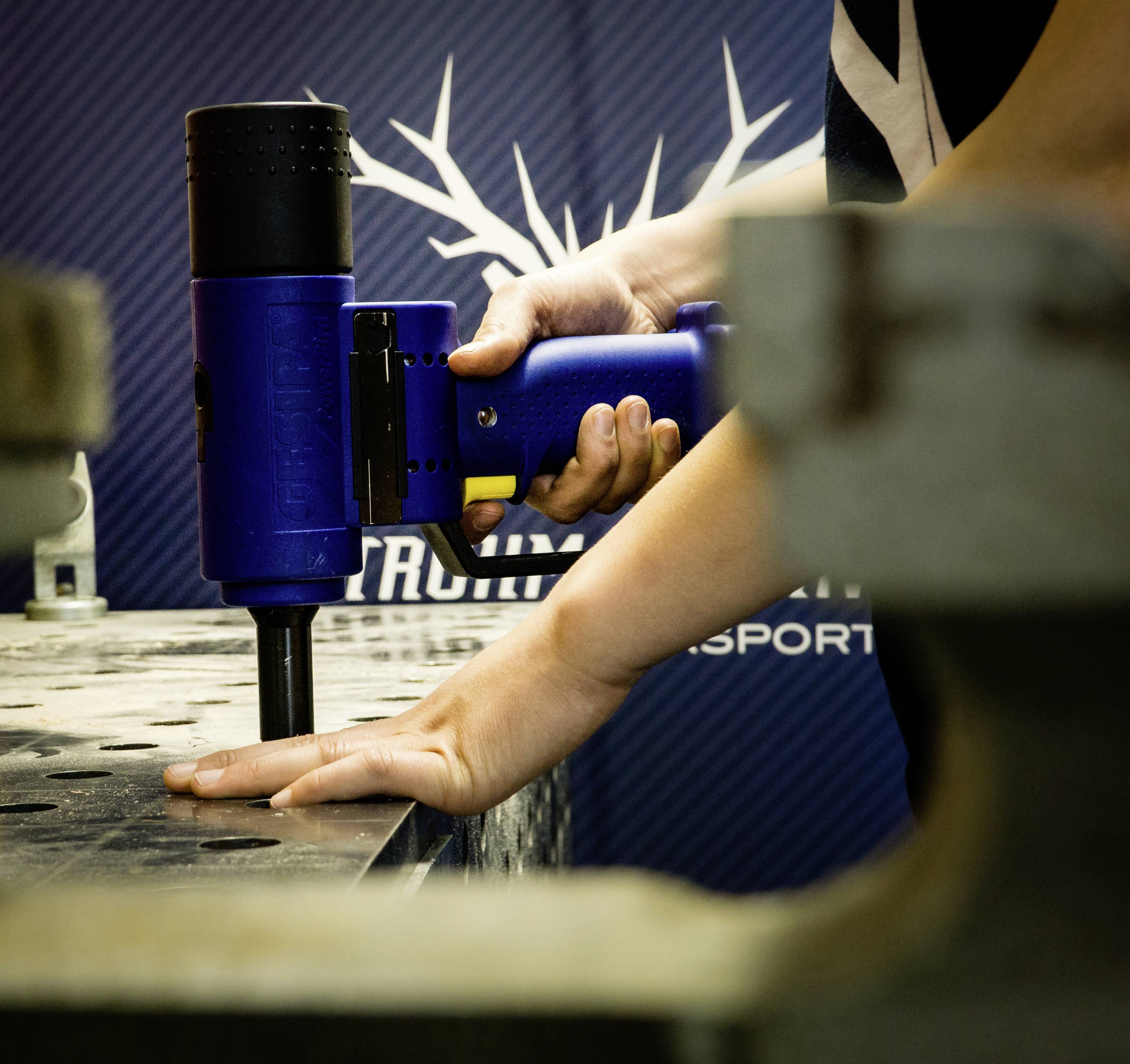 Osoba obsluhuje modrú vŕtačku na kovovej doske. V rozmazanom pozadí je firemné logo.