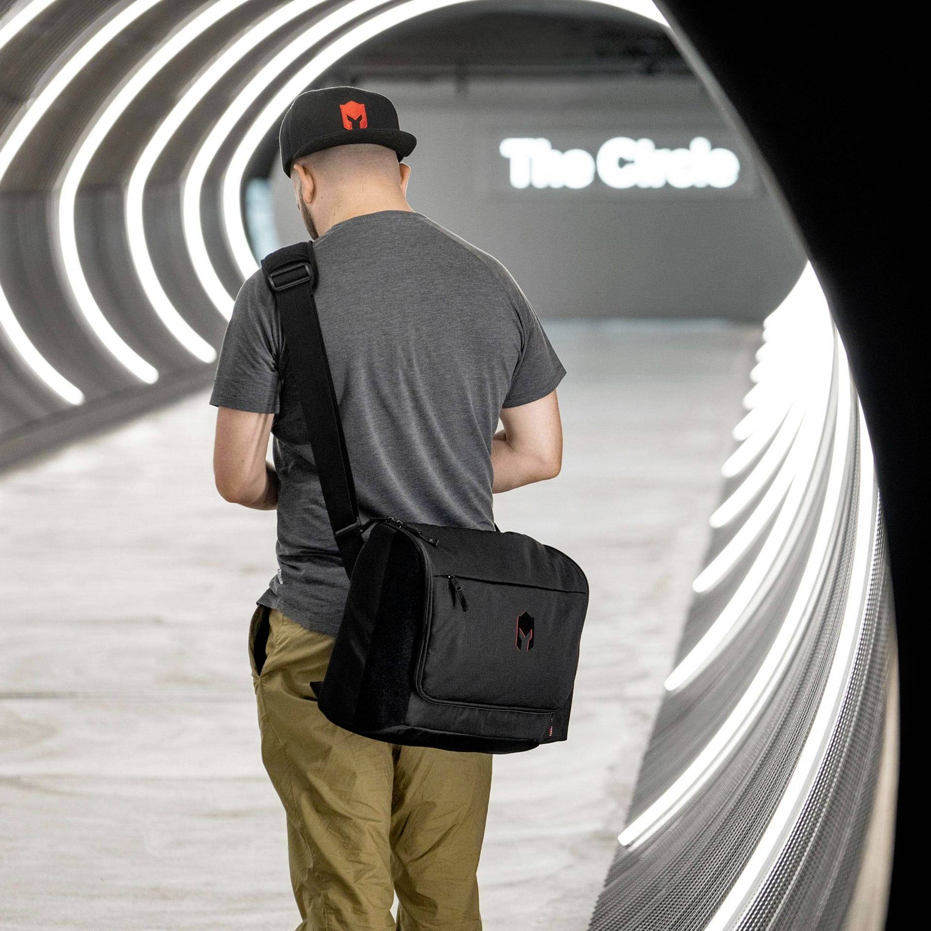 Muž s čiernou taškou a čiapkou kráča osvetleným tunelom. V pozadí je rozmazane viditeľný nápis 'The Circle'.